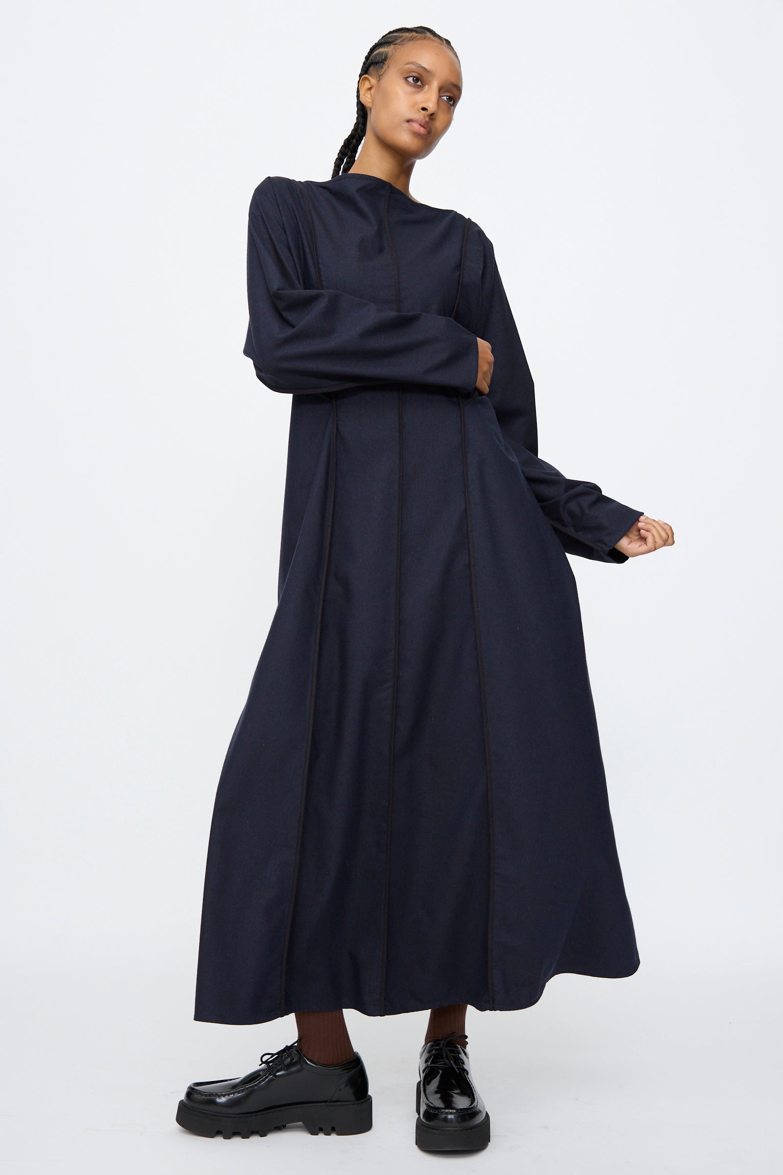 A person wearing the Baserange Nika Dress in Dama Navy—a navy, long sleeve, silk wool blend maxi dress with a boat neck—and black platform shoes stands against a plain white background.