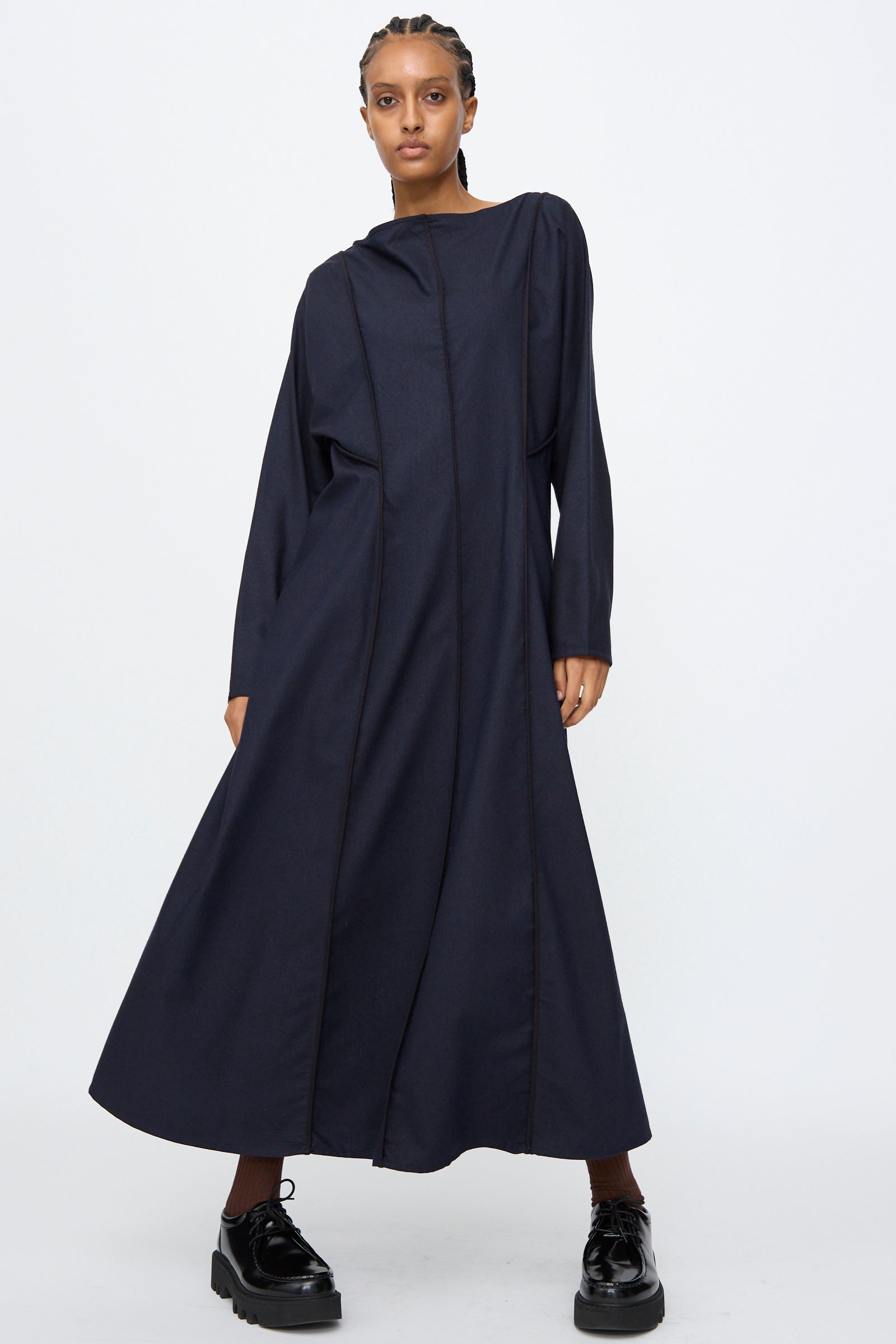 A person wears the Baserange Nika Dress in Dama Navy, a long sleeve maxi dress, paired with black platform shoes, standing against a plain white background.