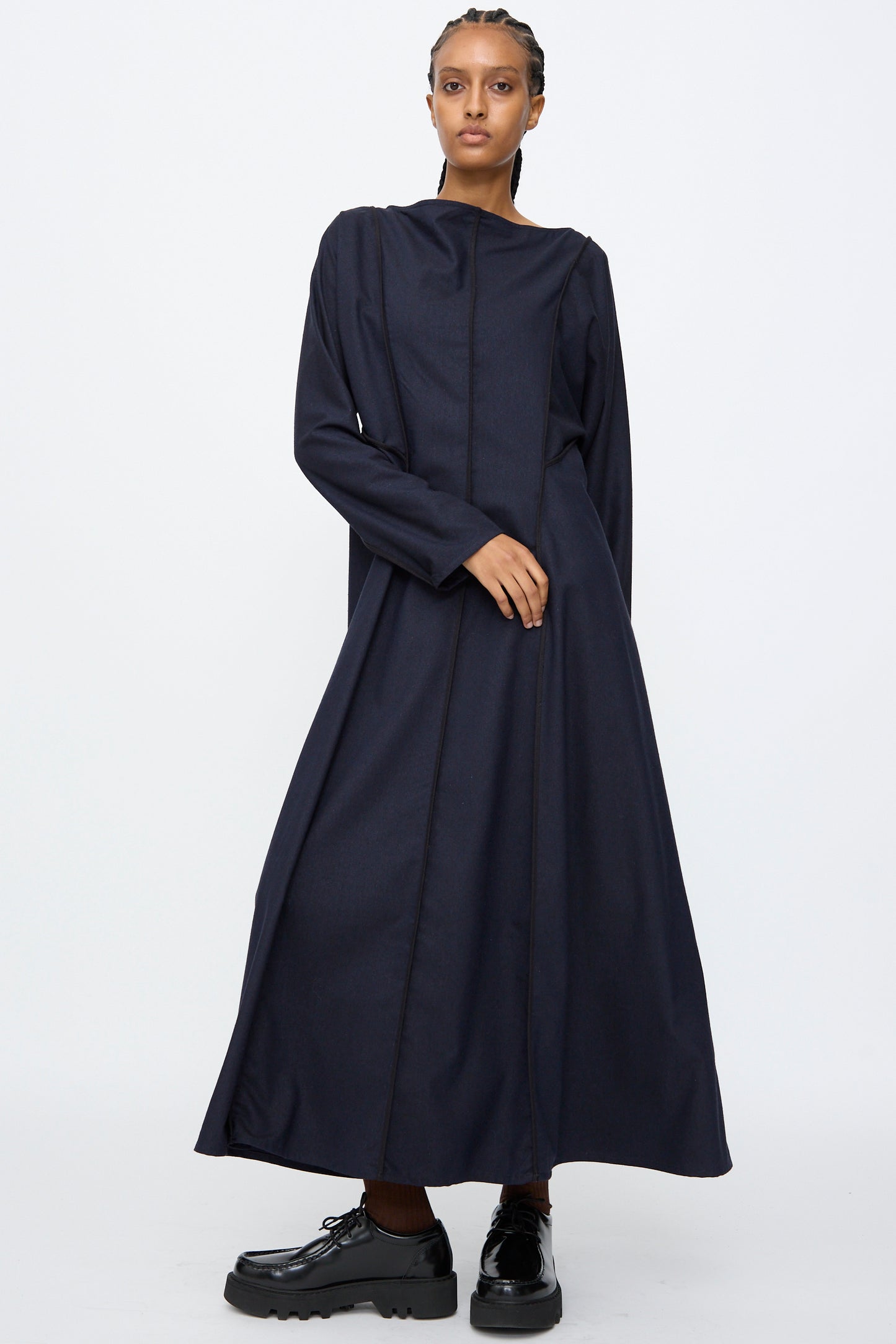 Model wears the Baserange Nika Dress in Dama Navy, a long sleeve maxi with a boat neck, paired with black platform shoes, standing against a plain white background.