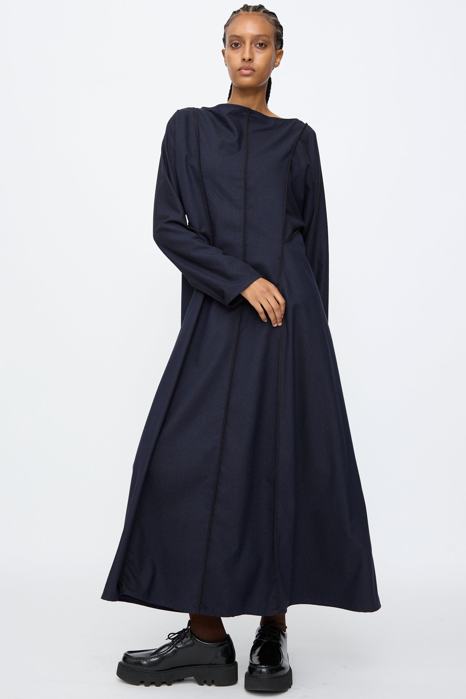 Model wears the Baserange Nika Dress in Dama Navy, a long sleeve maxi with a boat neck, paired with black platform shoes, standing against a plain white background.