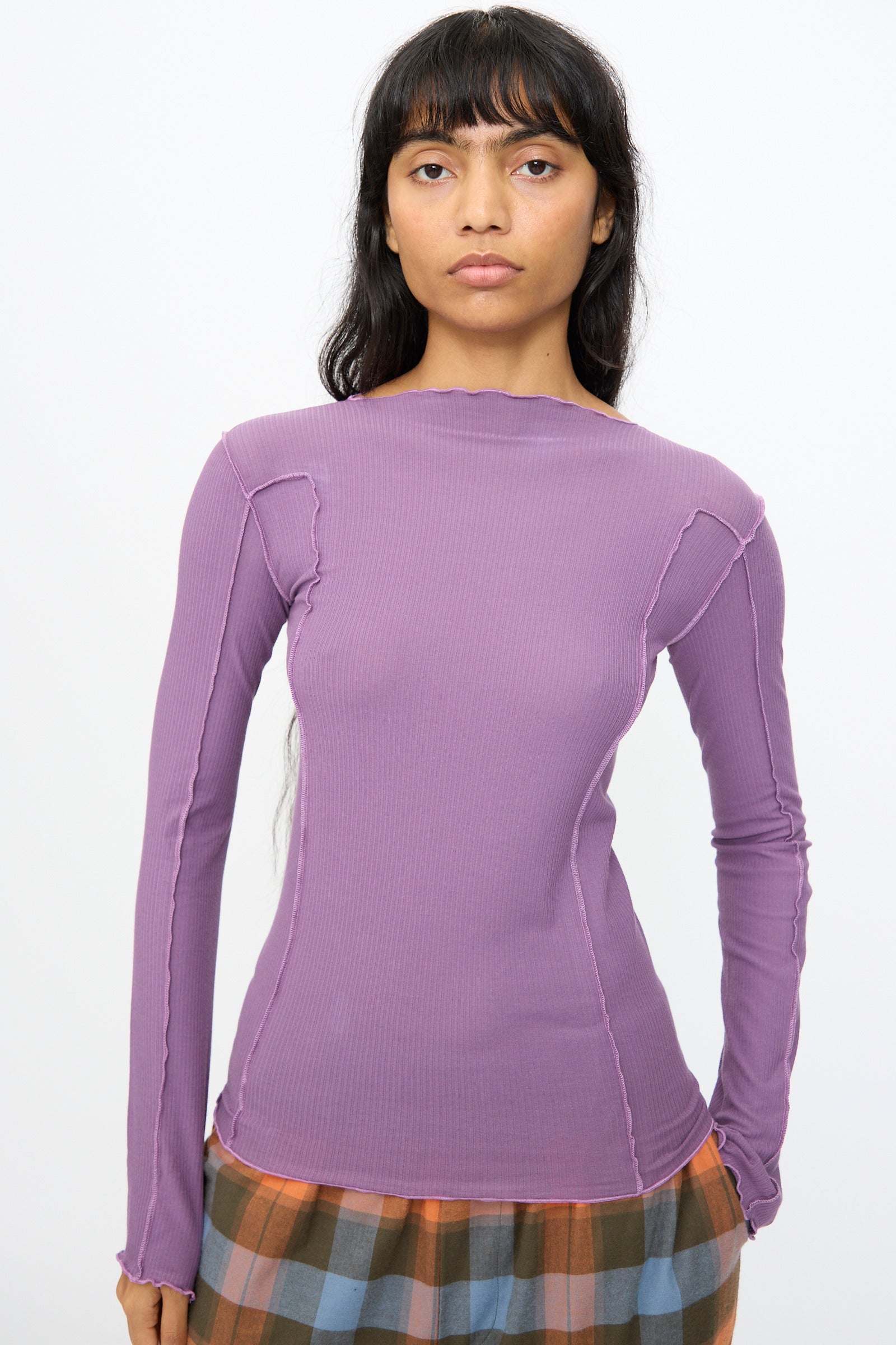 A woman wearing the Baserange Omato Long Sleeve Tee in Venet Purple stands against a plain white background, paired with a pleated, multi-colored checkered skirt.