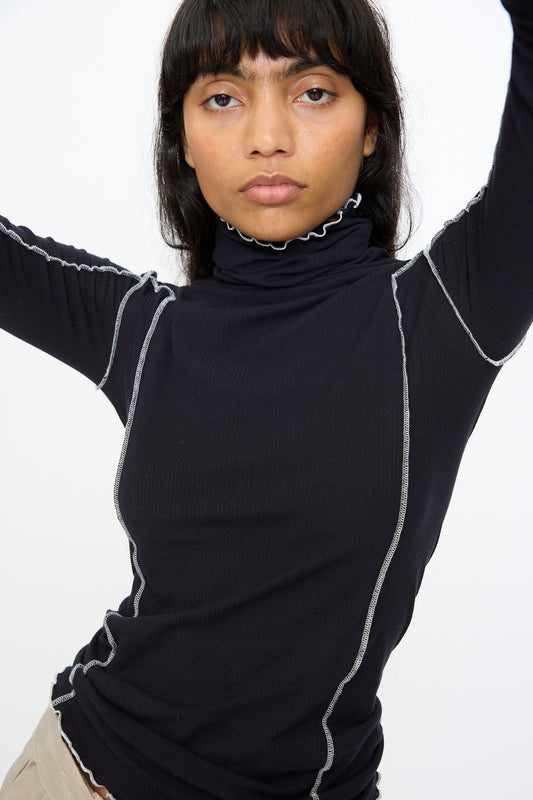 A person with long dark hair and bangs wears the Baserange Omato Turtleneck in Murano Navy and Undyed, featuring ribbed organic cotton and contrast exposed seams, posing with arms raised against a plain white background.