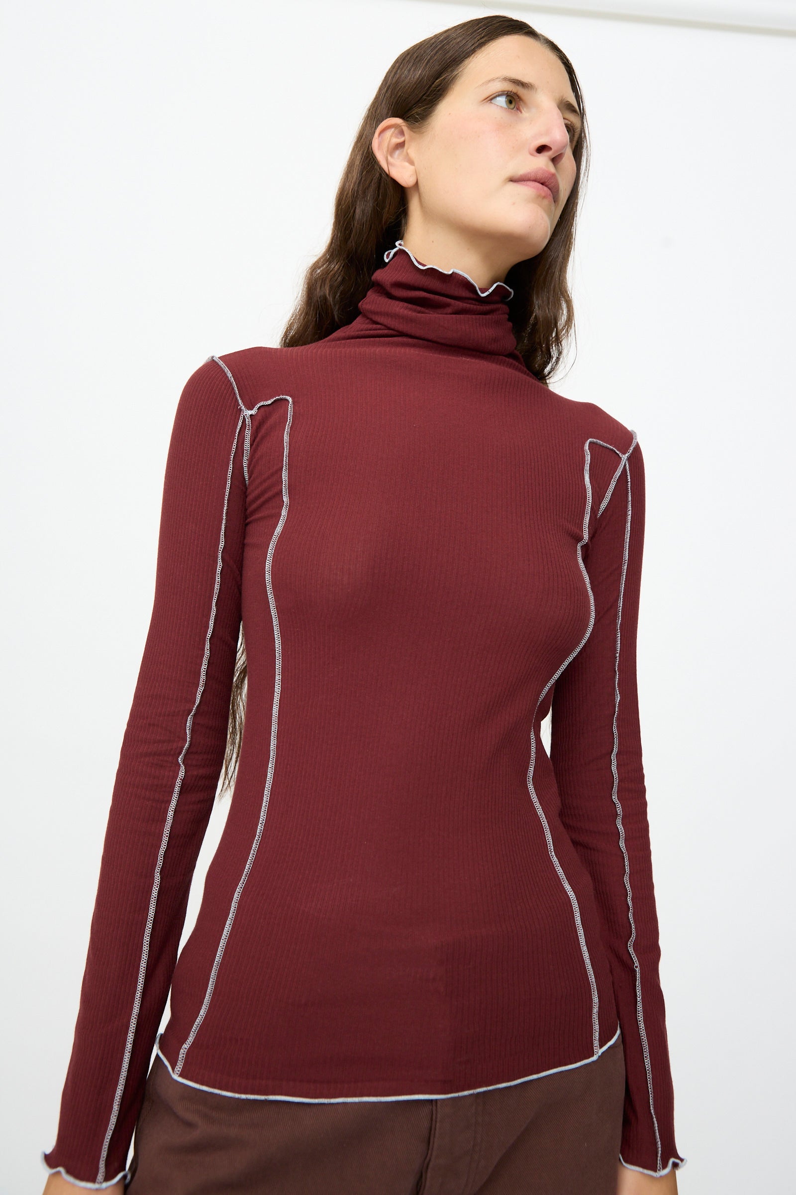 A person stands against a plain white background wearing the Baserange Omato Turtleneck in Nara Red, a women’s slim-fit long-sleeve top featuring white contrast stitching.