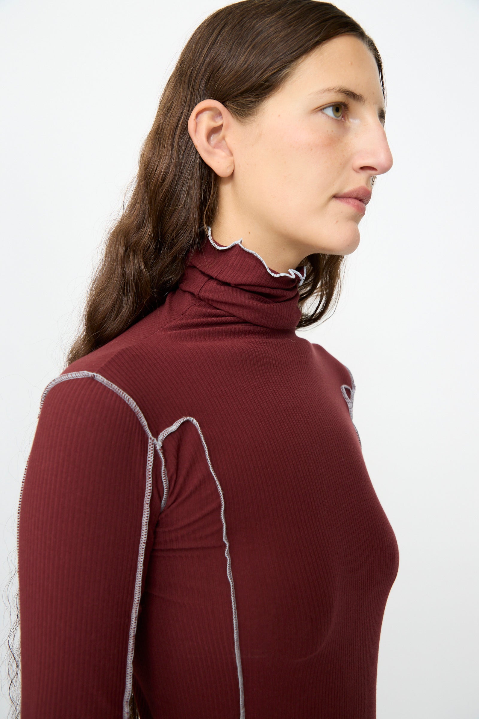 A person with long brown hair is wearing the Baserange Omato Turtleneck in Nara Red, styled in a women’s slim fit with white stitching, and is looking to the right against a plain white background.