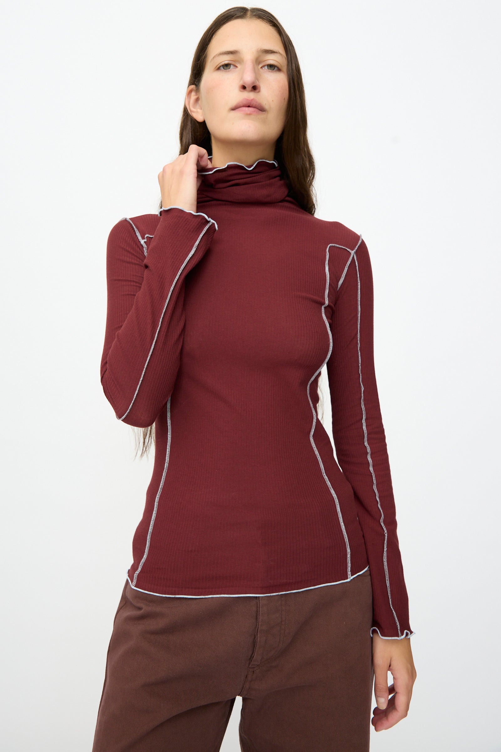 A person is wearing the Baserange Omato Turtleneck in Nara Red, featuring white seam details, paired with brown pants and standing against a plain white background.