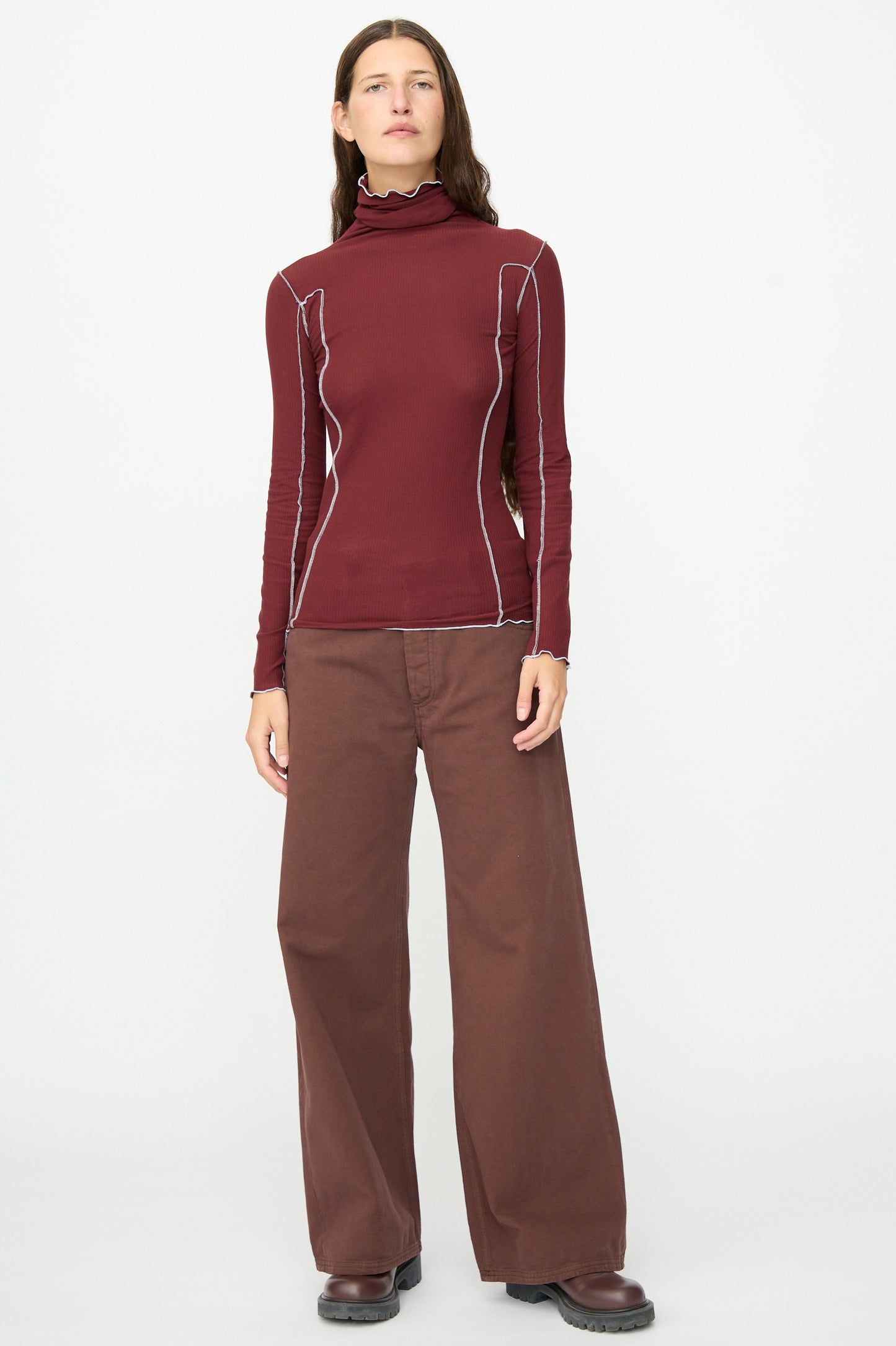 A person stands facing forward, wearing the Baserange Omato Turtleneck in Nara Red, styled with wide brown pants and dark shoes against a plain white background.