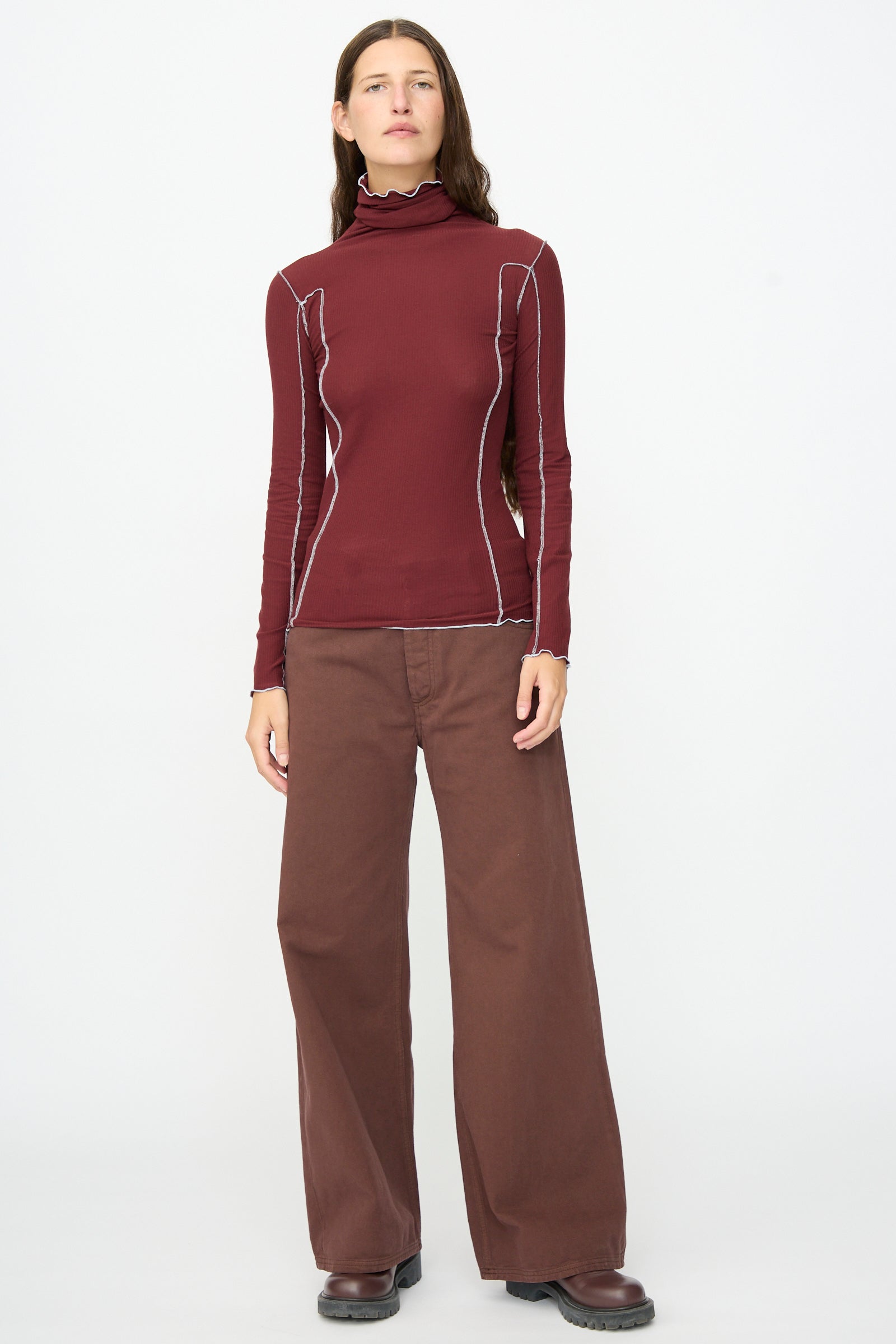 A person stands facing forward, wearing the Baserange Omato Turtleneck in Nara Red, styled with wide brown pants and dark shoes against a plain white background.