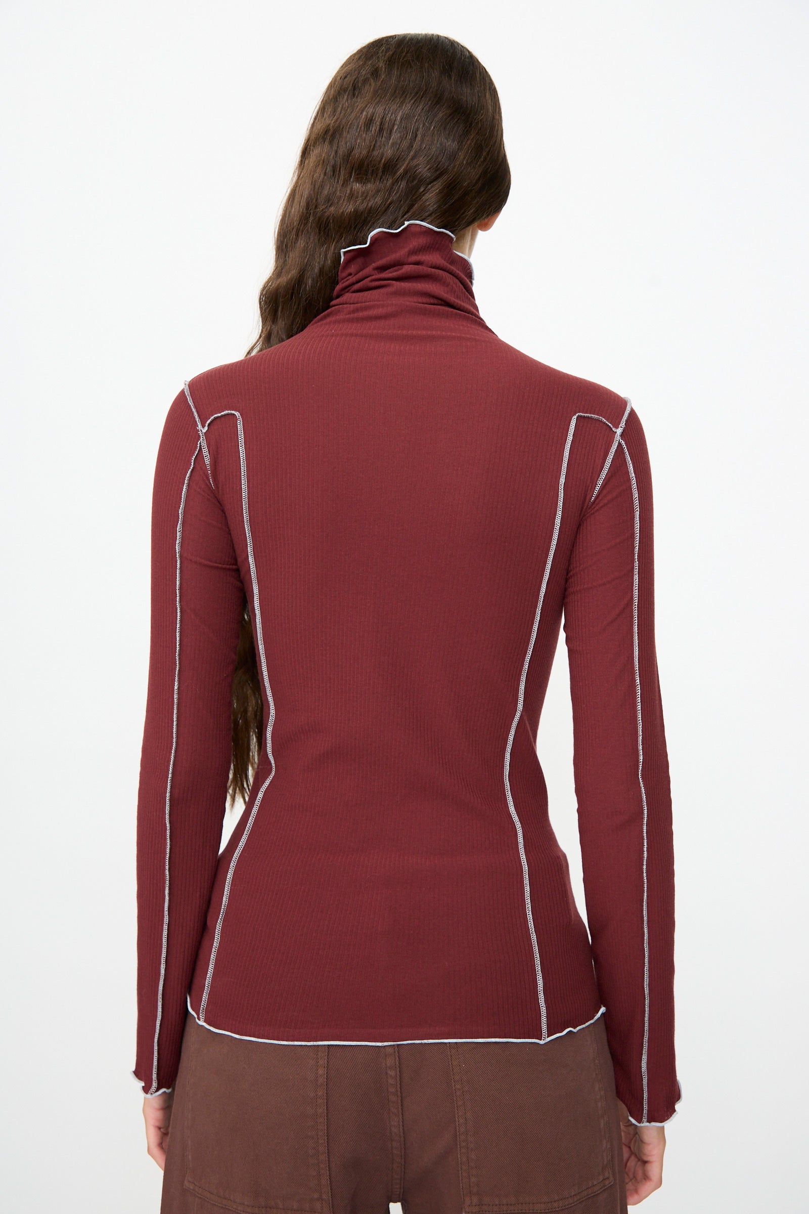 A person with long brown hair is shown from the back, wearing the Baserange Omato Turtleneck in Nara Red—a women’s slim fit, long sleeve top with white stitching—against a plain background.
