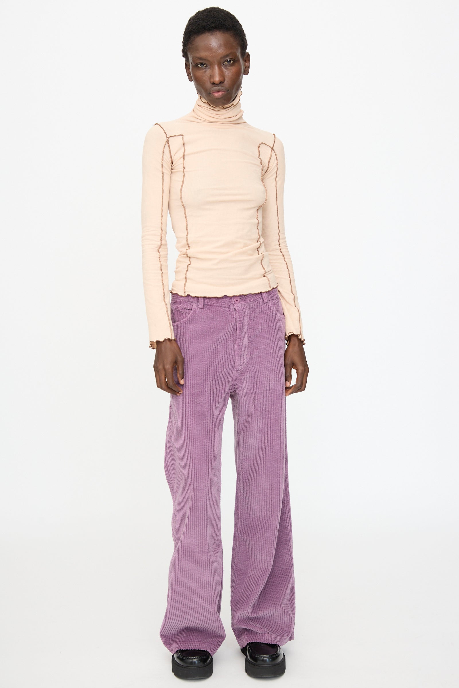 A person stands against a white background wearing Baserange's Omato Turtleneck in Tin Beige and Ocular, paired with loose purple corduroy pants and black shoes.