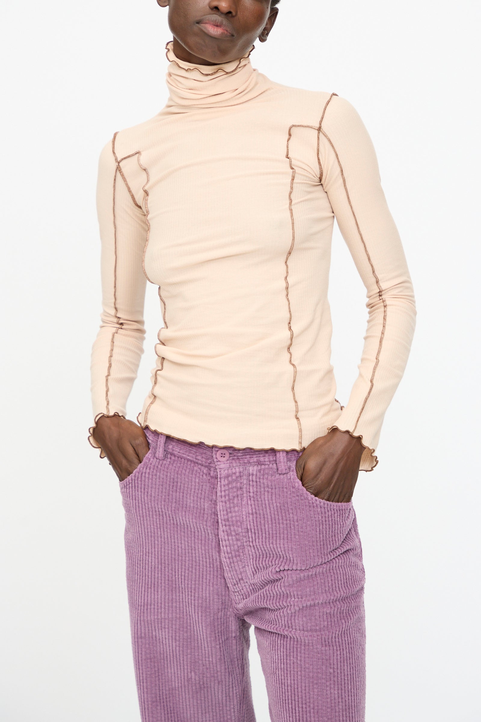 Model wears the Baserange Omato Turtleneck in Tin Beige and Ocular, a ribbed organic cotton top with exposed seams, paired with high-waisted lavender corduroy pants, hands in pockets against a plain white background.
