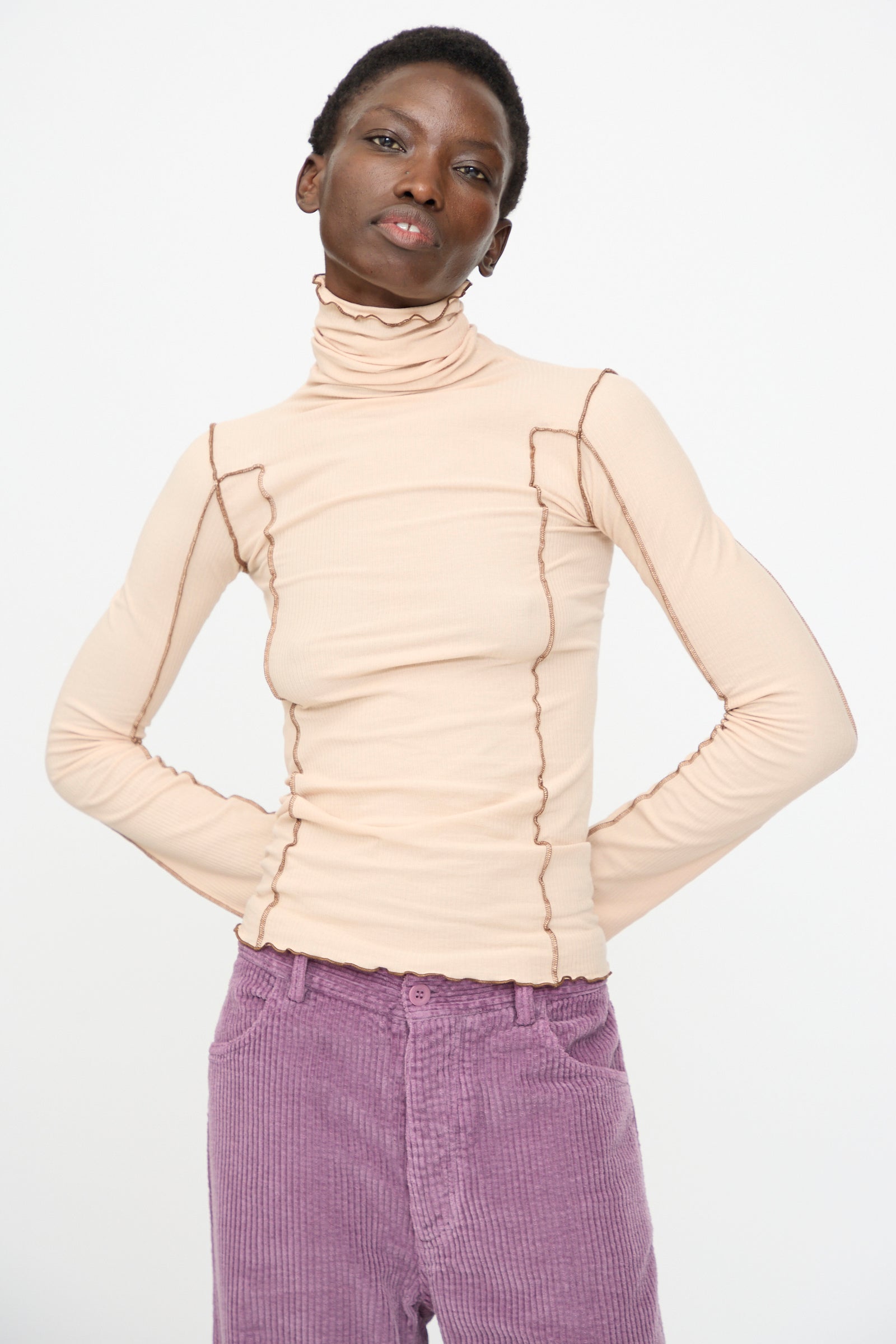 Model wears the Baserange Omato Turtleneck in Tin Beige with exposed seams and purple corduroy pants, standing hands on hips against a plain white background.