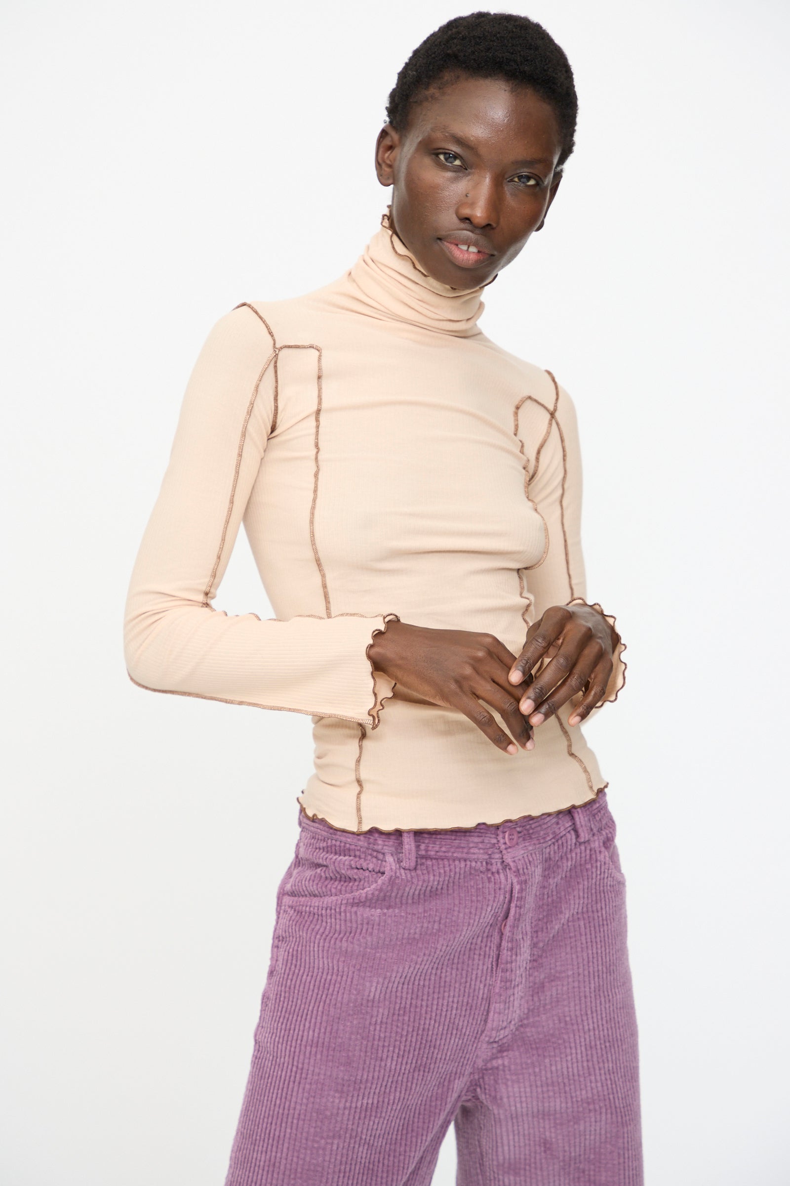 A person wears the Baserange Omato Turtleneck in Tin Beige and Ocular, paired with lilac corduroy pants, standing against a plain white background.