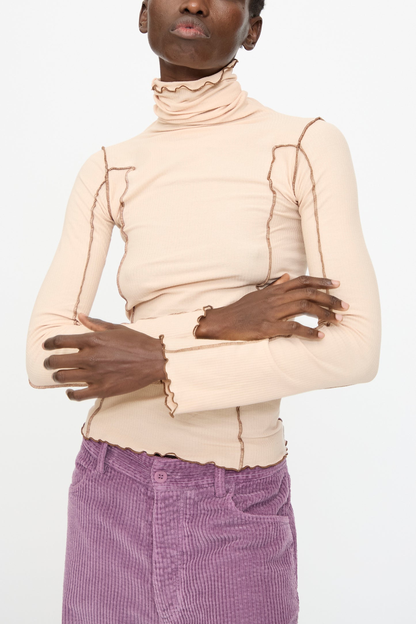 A person stands with arms crossed against a plain background, wearing the Baserange Omato Turtleneck in Tin Beige and Ocular, styled with light purple corduroy pants.