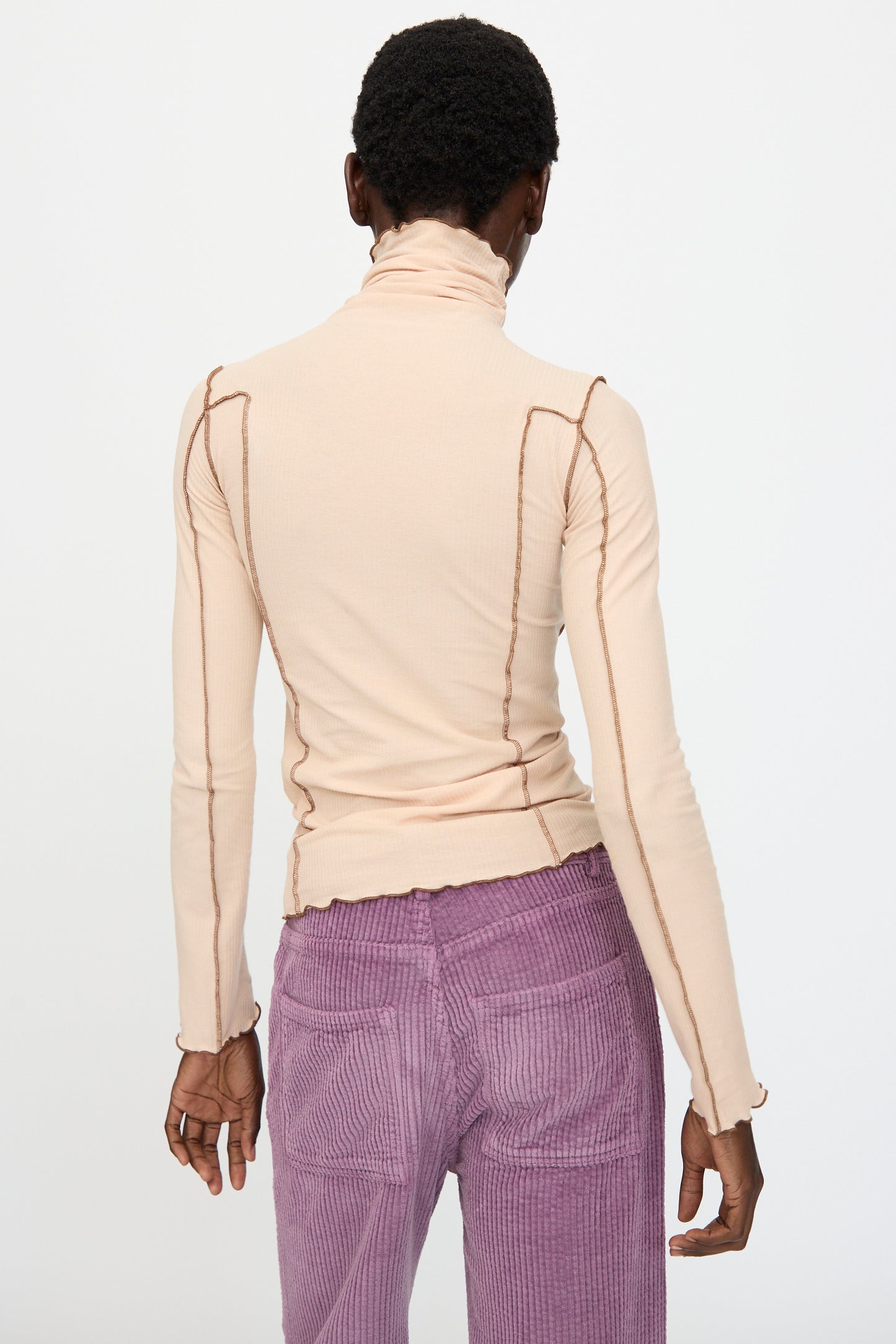 A person stands with their back to the camera, wearing the Baserange Omato Turtleneck in Tin Beige and Ocular, paired with high-waisted purple corduroy pants.