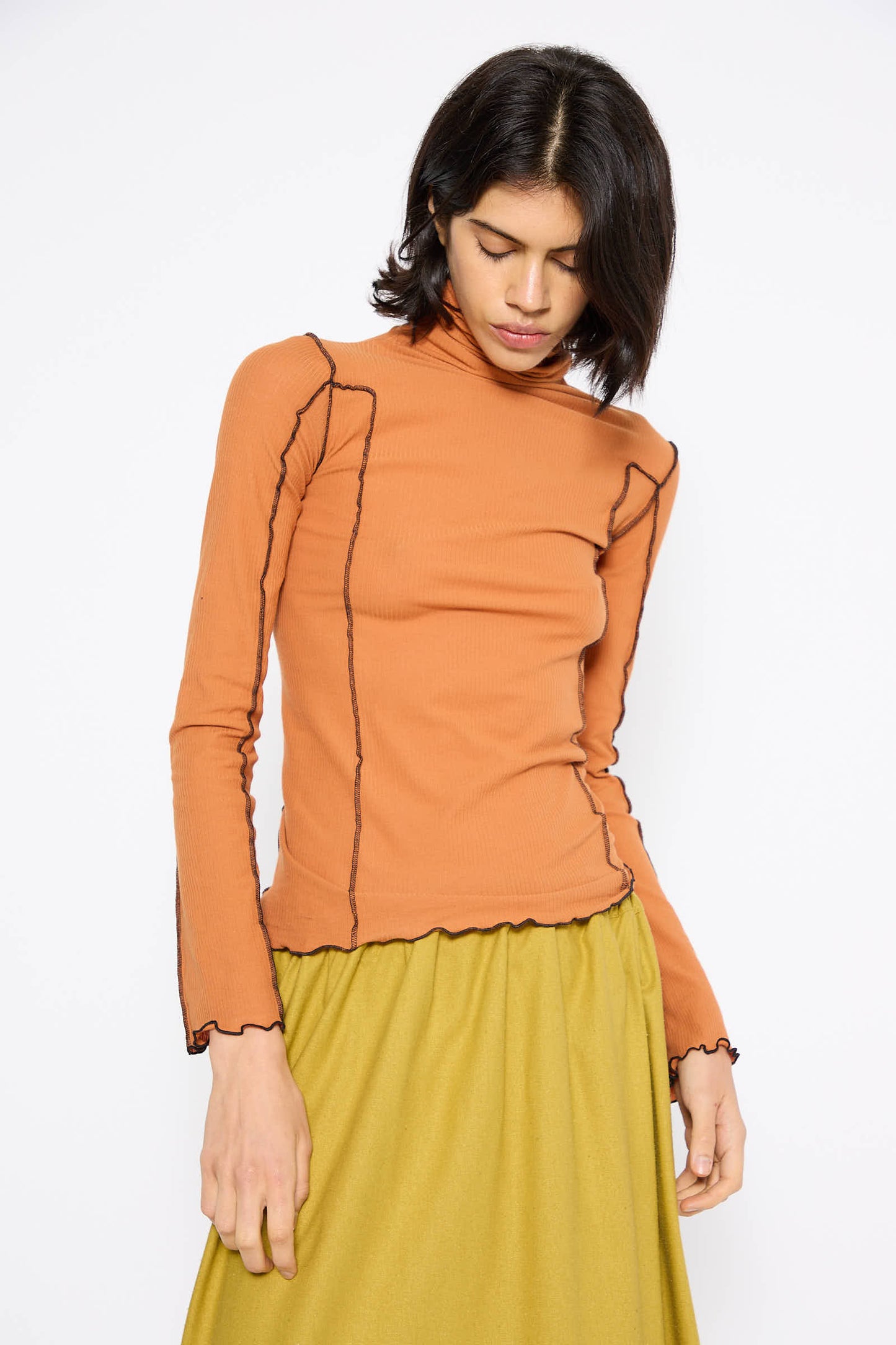 A person stands wearing the Omato Turtleneck in Dre Orange by Baserange, a long-sleeve ribbed knit tee with black seam detailing, paired with a yellow skirt. The background is plain white.