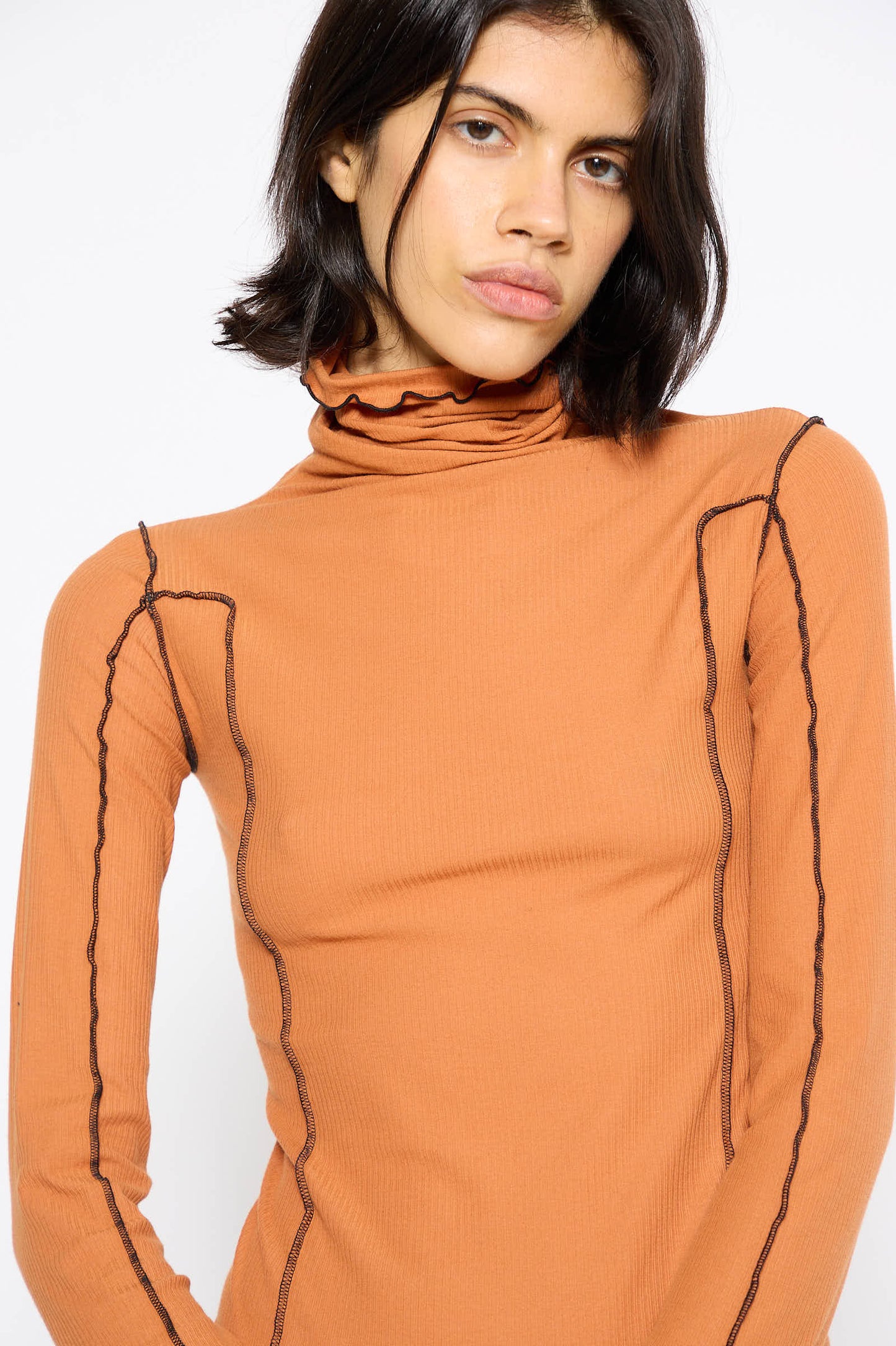 A woman with shoulder-length dark hair, wearing the Baserange Omato Turtleneck in Dre Orange, stands against a plain white background.