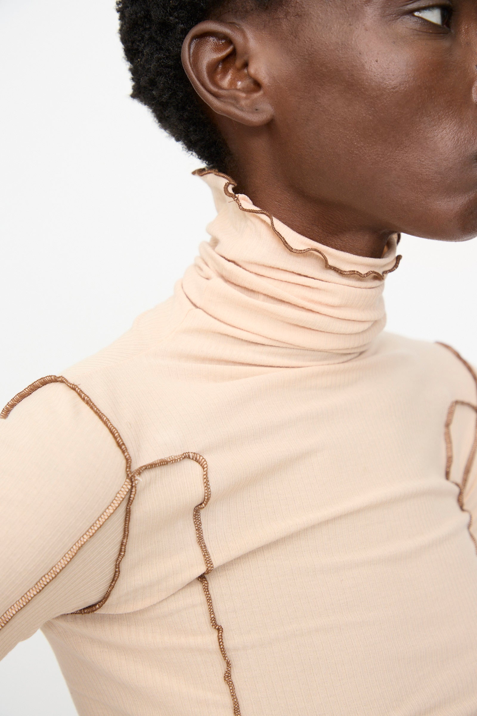 A person wears the Baserange Omato Turtleneck in Tin Beige and Ocular, a ribbed organic cotton top with brown exposed seams, shown from the shoulders up against a plain background.
