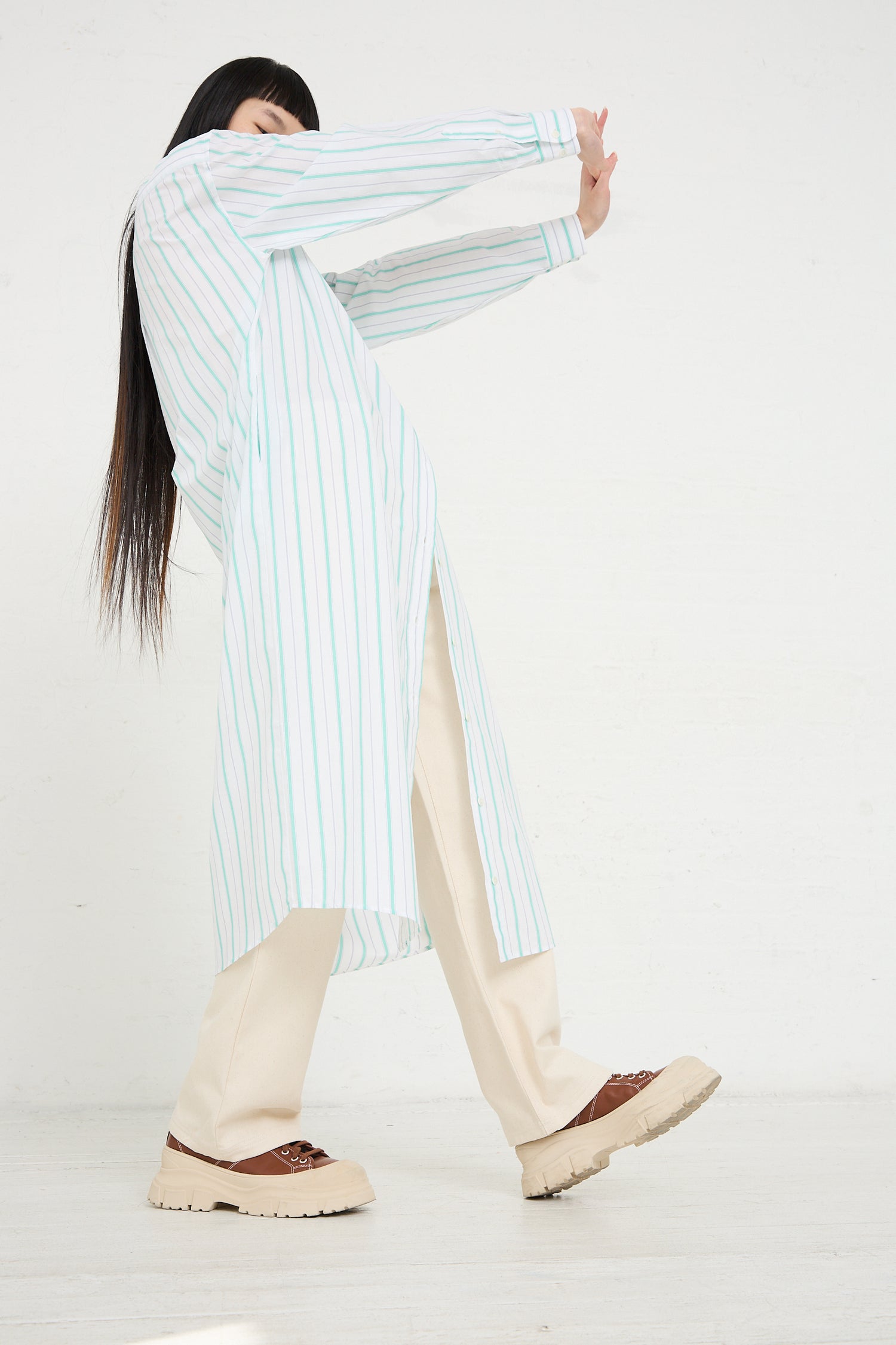 A woman poses sideways in a Baserange Organic Cotton Ole Shirt Dress in Stripe and cream trousers, with her face obscured by her arm.