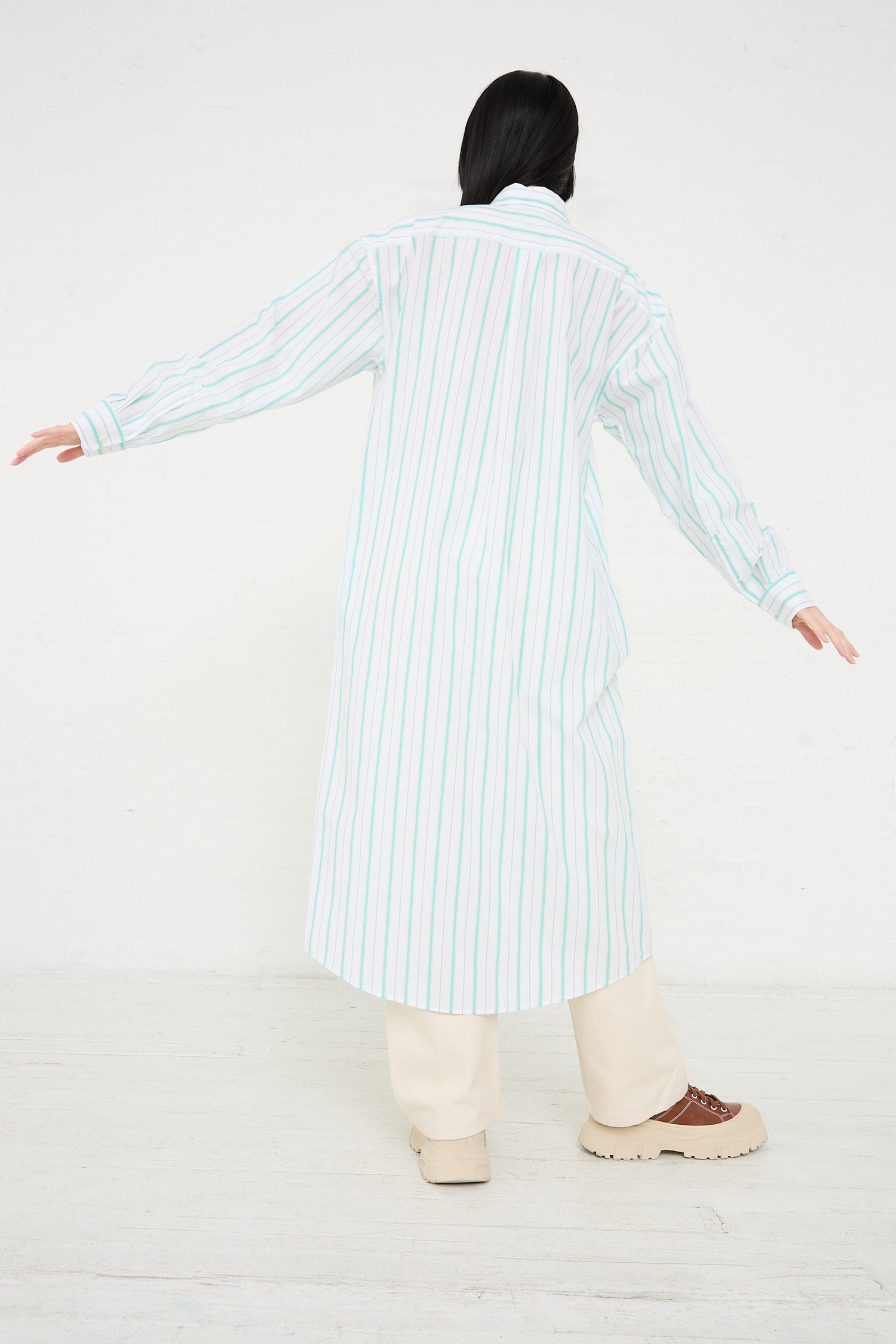 A person viewed from behind, wearing a Baserange Organic Cotton Ole Shirt Dress in Stripe and cream pants, with arms outstretched, standing against a white background.