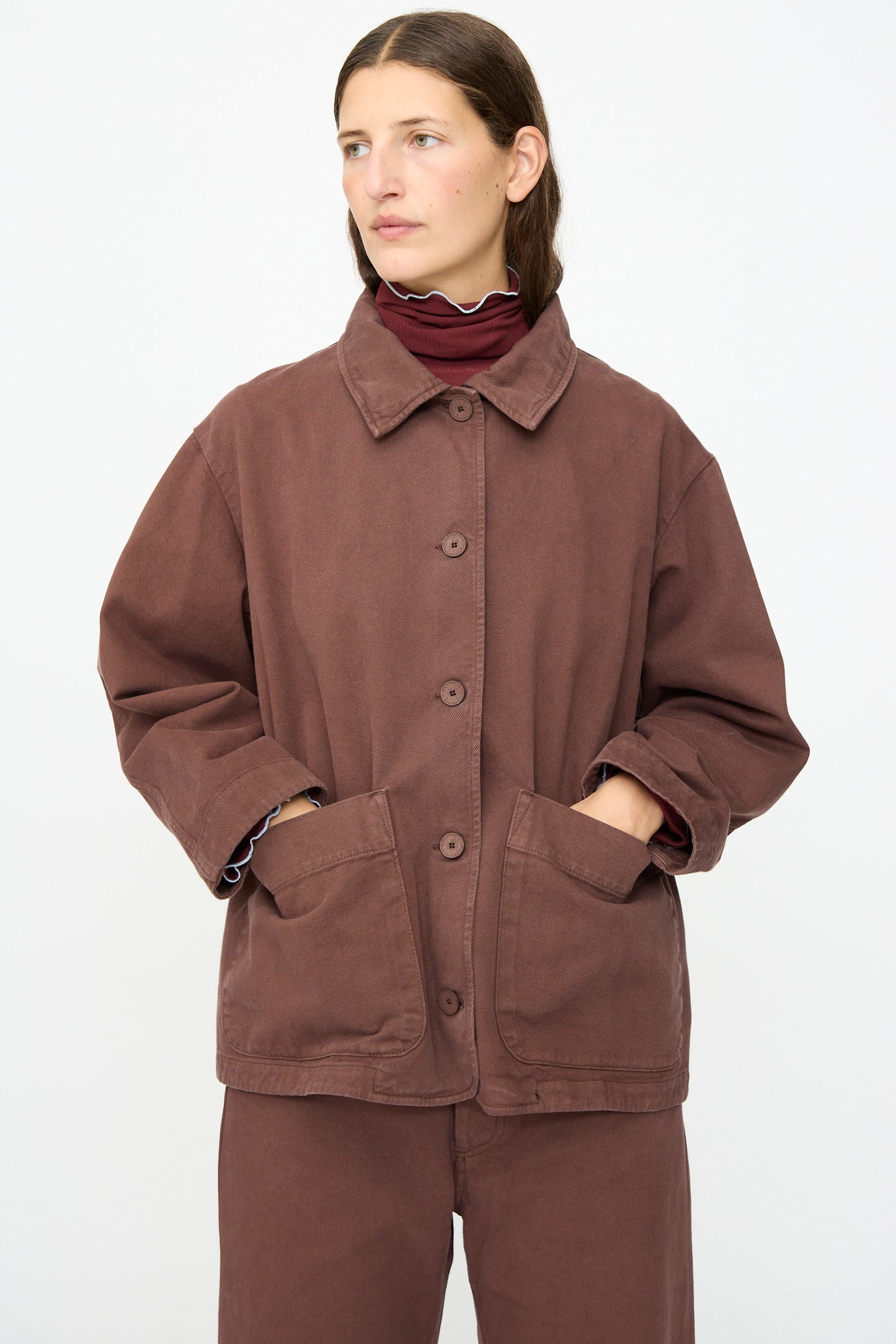 Wearing the Baserange Osi Jacket in Cata Brown—a relaxed gabardine cotton piece with large front pockets, made in Portugal—this person stands against a plain white background.