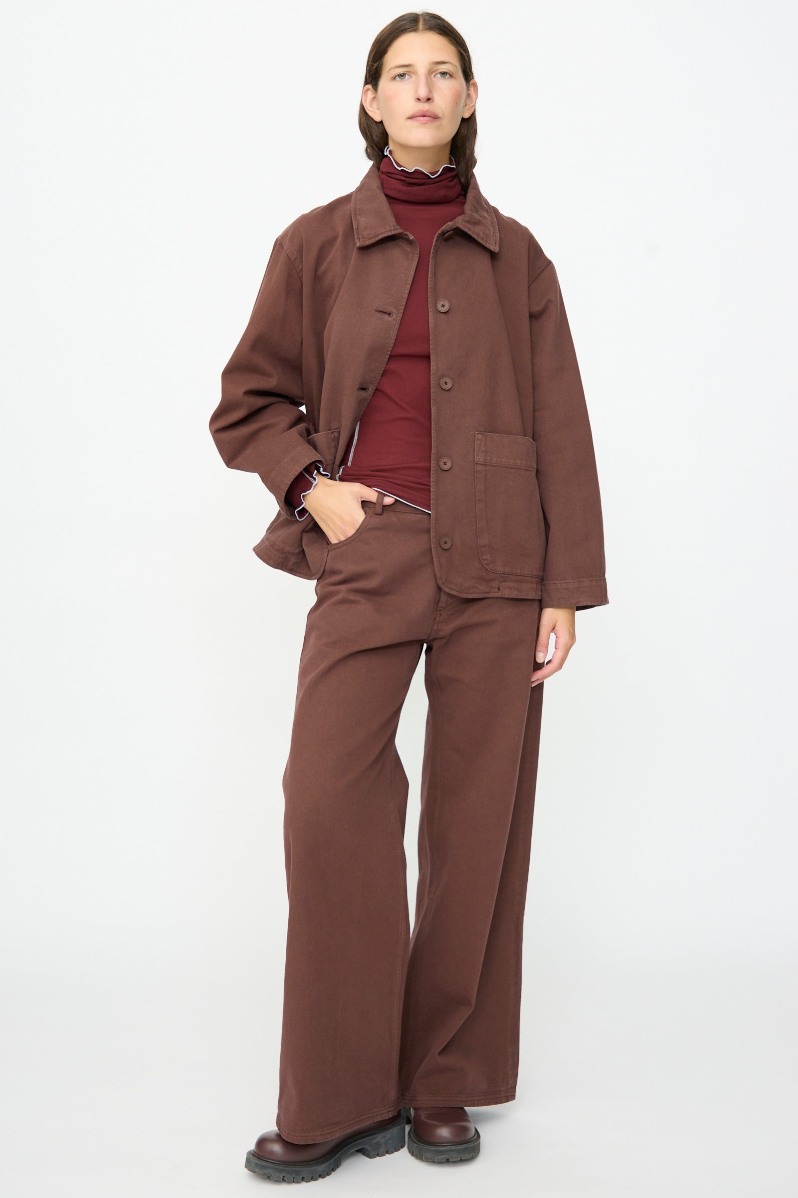 A person stands against a plain white background wearing a brown wide-leg pantsuit, burgundy turtleneck, and black shoes, topped with the Baserange Osi Jacket in Cata Brown—a relaxed gabardine cotton jacket made in Portugal.