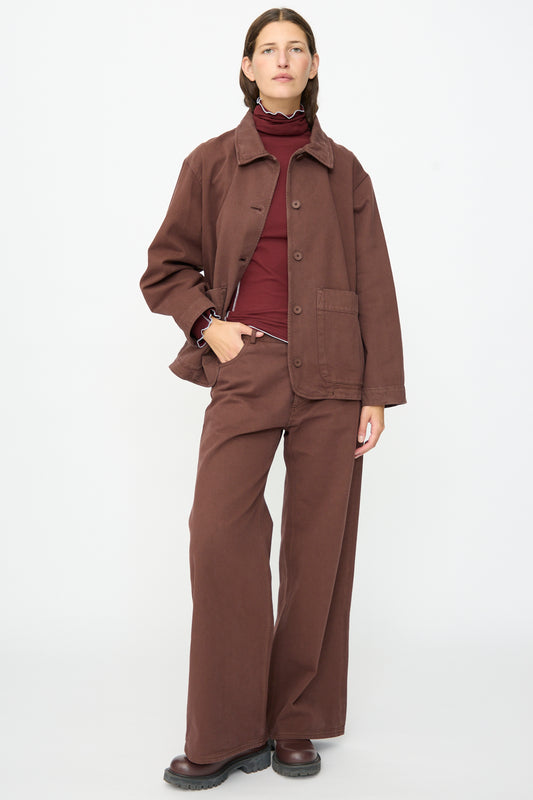 A person stands against a plain white background wearing a brown wide-leg pantsuit, burgundy turtleneck, and black shoes, topped with the Baserange Osi Jacket in Cata Brown—a relaxed gabardine cotton jacket made in Portugal.