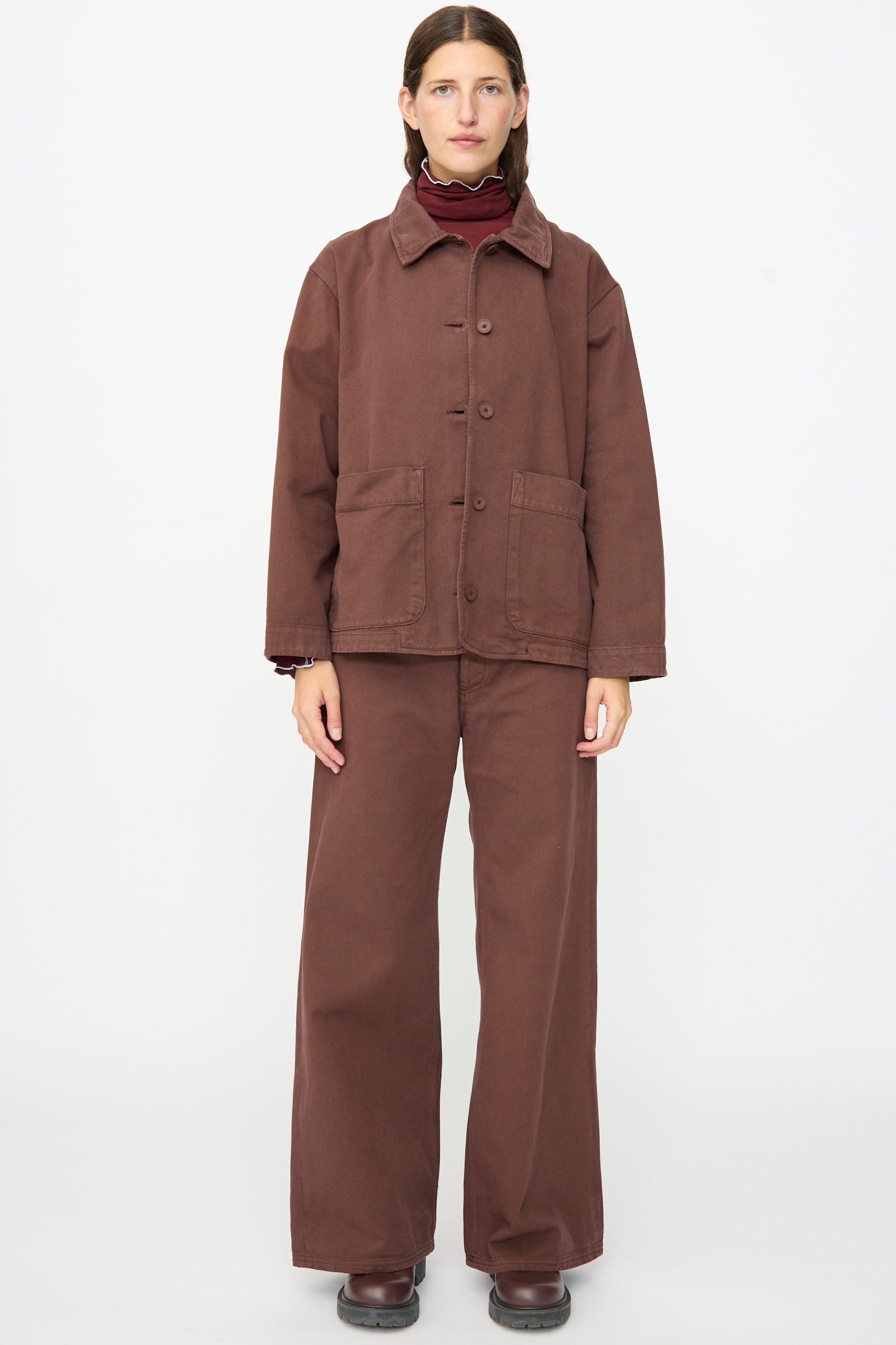 A person stands facing forward against a plain white background, wearing the Baserange Osi Jacket in Cata Brown—a relaxed gabardine cotton jacket made in Portugal—paired with matching wide-leg pants, a maroon turtleneck sweater, and brown shoes.