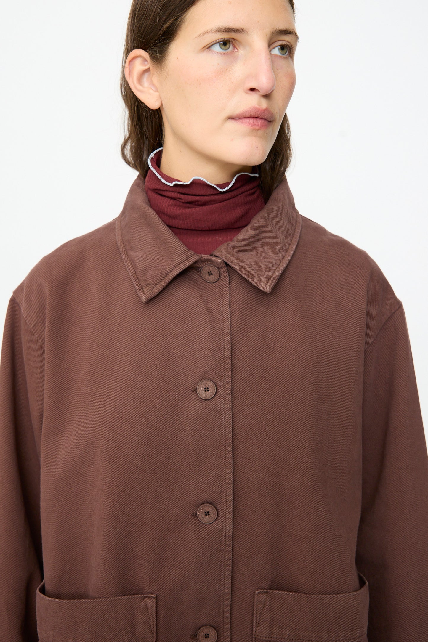 A person wearing the Baserange Osi Jacket in Cata Brown, crafted from gabardine cotton in Portugal, over a maroon turtleneck, gazes slightly to the side against a plain background.