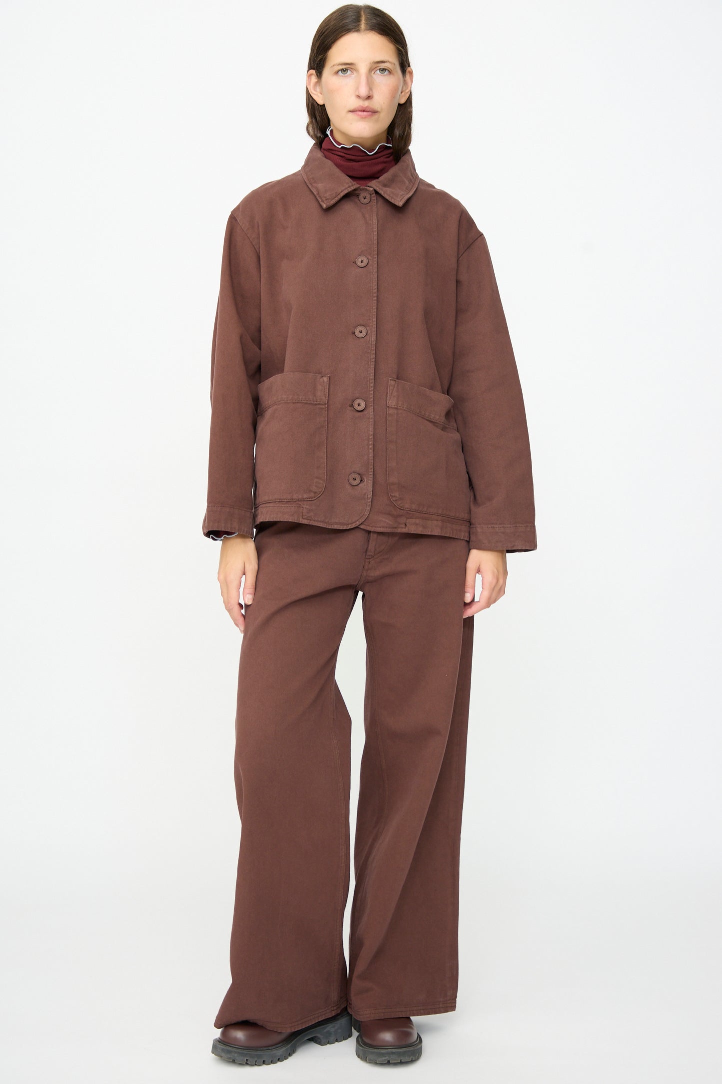 A person stands in a studio setting, wearing the Baserange Osi Jacket in Cata Brown—crafted from gabardine cotton in Portugal—with matching wide-leg pants and dark shoes, their straight hair and neutral expression completing the look.