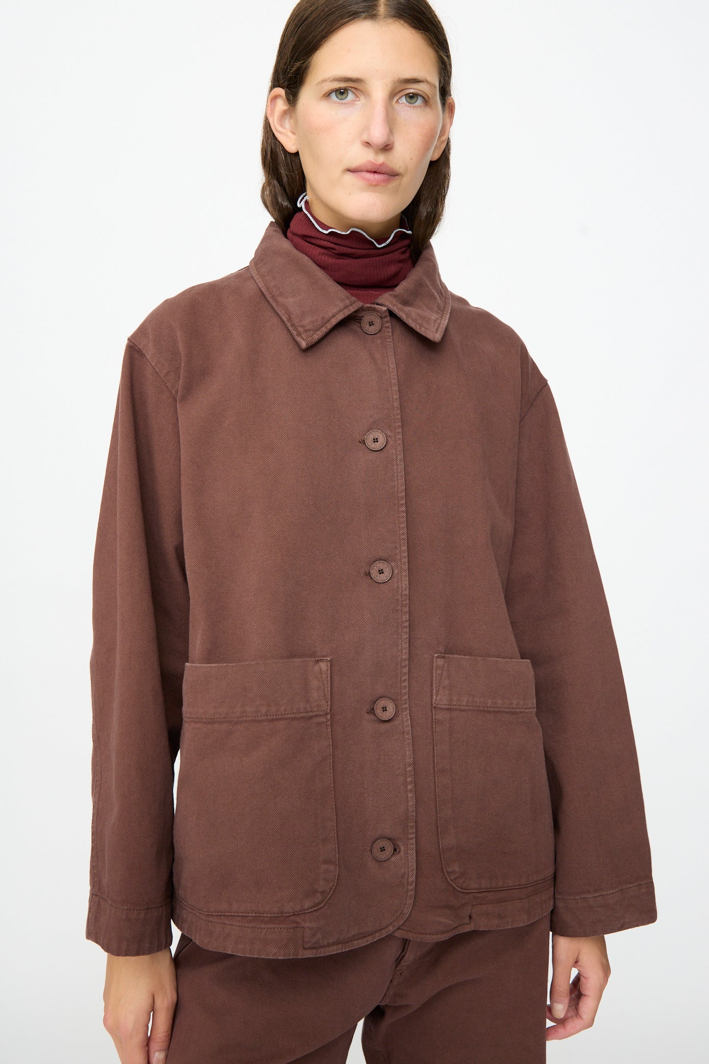 A person is wearing the Baserange Osi Jacket in Cata Brown, a relaxed gabardine cotton piece made in Portugal, featuring two front pockets and paired with matching brown pants, while standing against a plain white background.