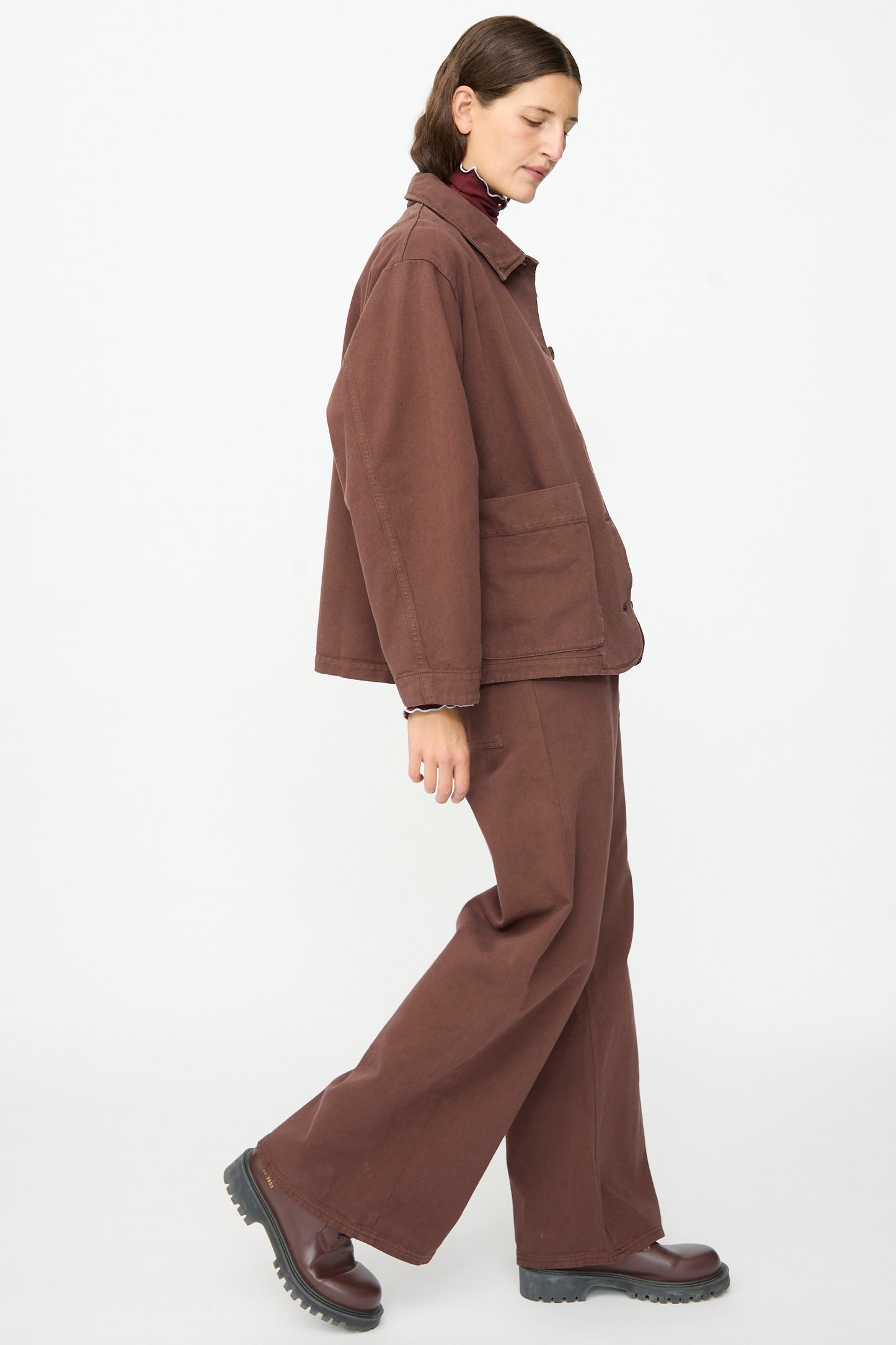 A person walks to the left against a plain white background, wearing Baserange’s Osi Jacket in Cata Brown with matching wide-leg pants and dark brown shoes.