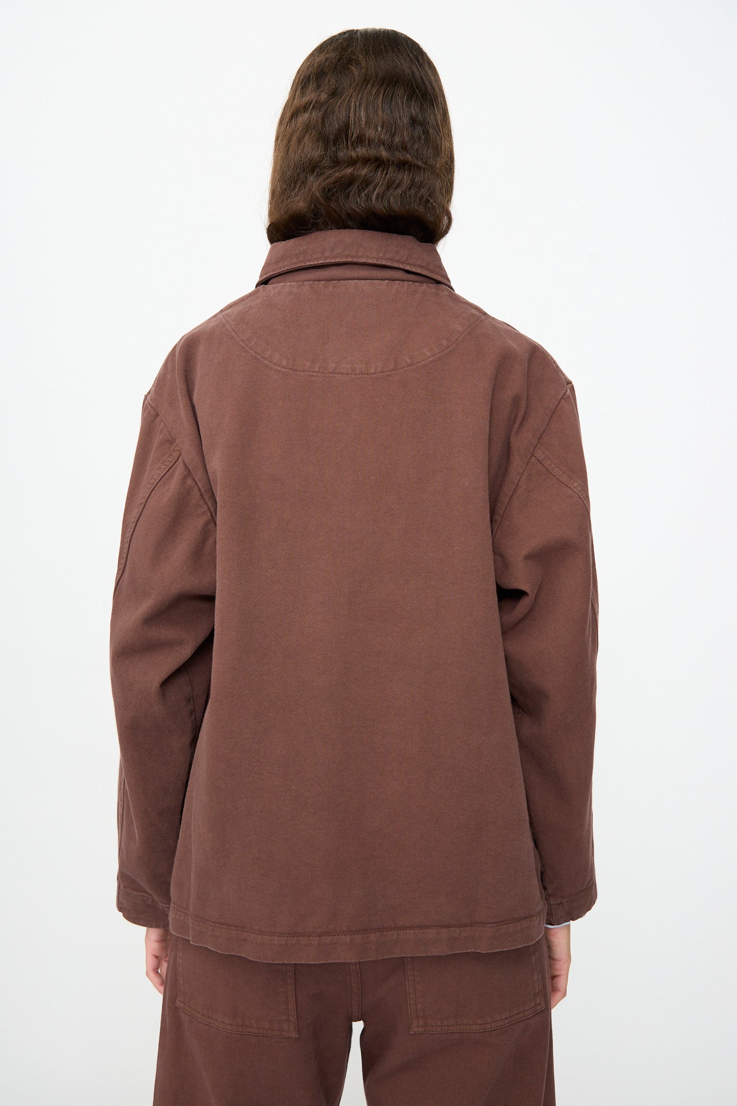A person stands with their back to the camera, dressed in the Baserange Osi Jacket in Cata Brown—crafted from relaxed gabardine cotton and made in Portugal—with matching pants, set against a plain light background.