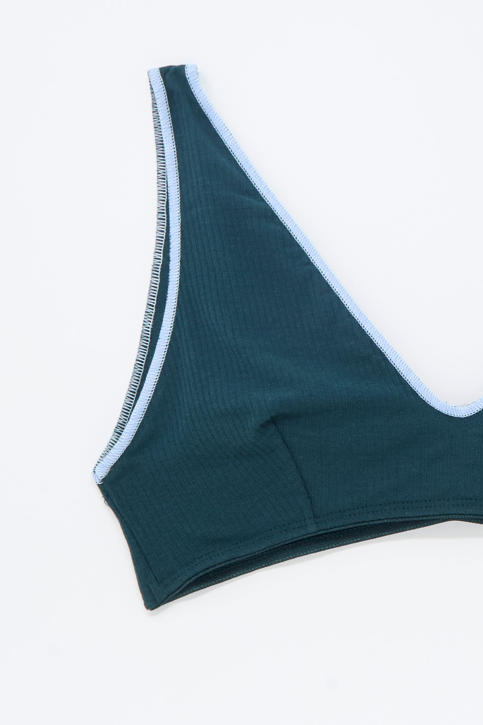 A close-up of the Baserange Pam Bra in Hirsu Green, featuring teal ribbed fabric with light blue trim and a V-neck design, laid flat on a white background.