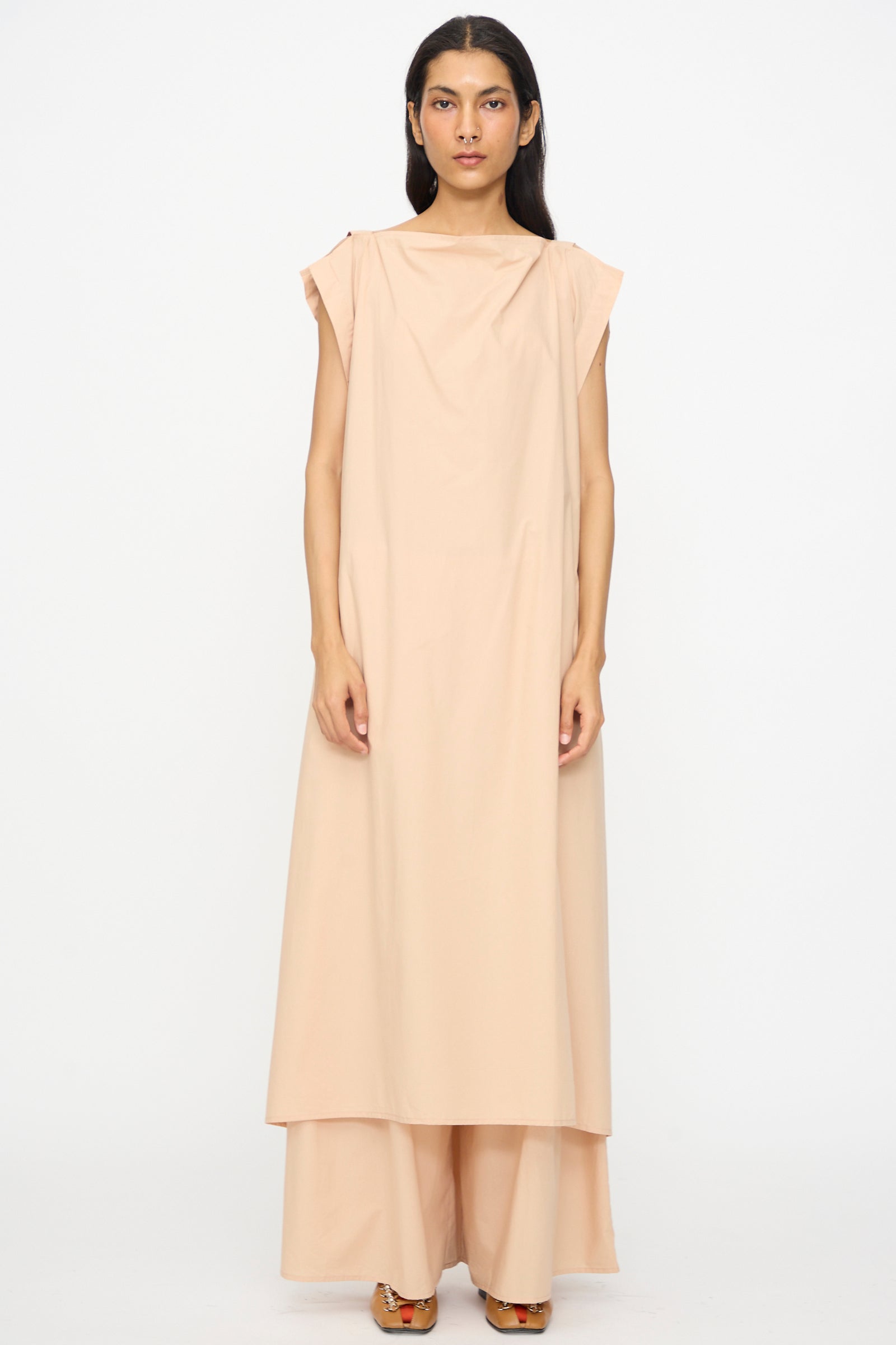 A woman faces forward wearing the Baserange Part Dress in Andradita Sand—an oversized, sleeveless light beige cotton poplin dress paired with wide-leg pants and brown shoes against a plain white background.