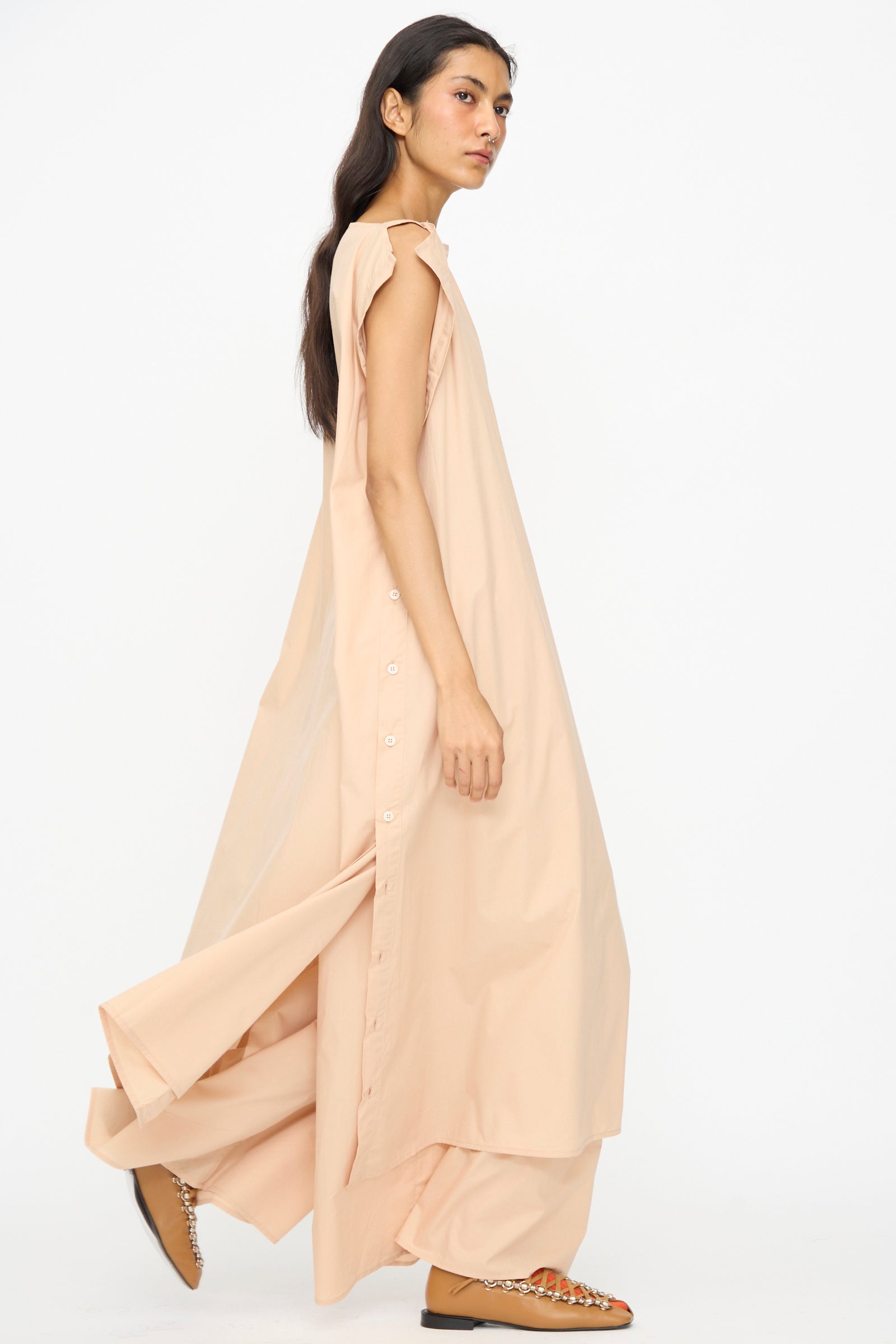 A woman wearing the Baserange Part Dress in Andradita Sand—an oversized, sleeveless, light peach midi dress with side buttons and wide pants—walks against a plain white background.