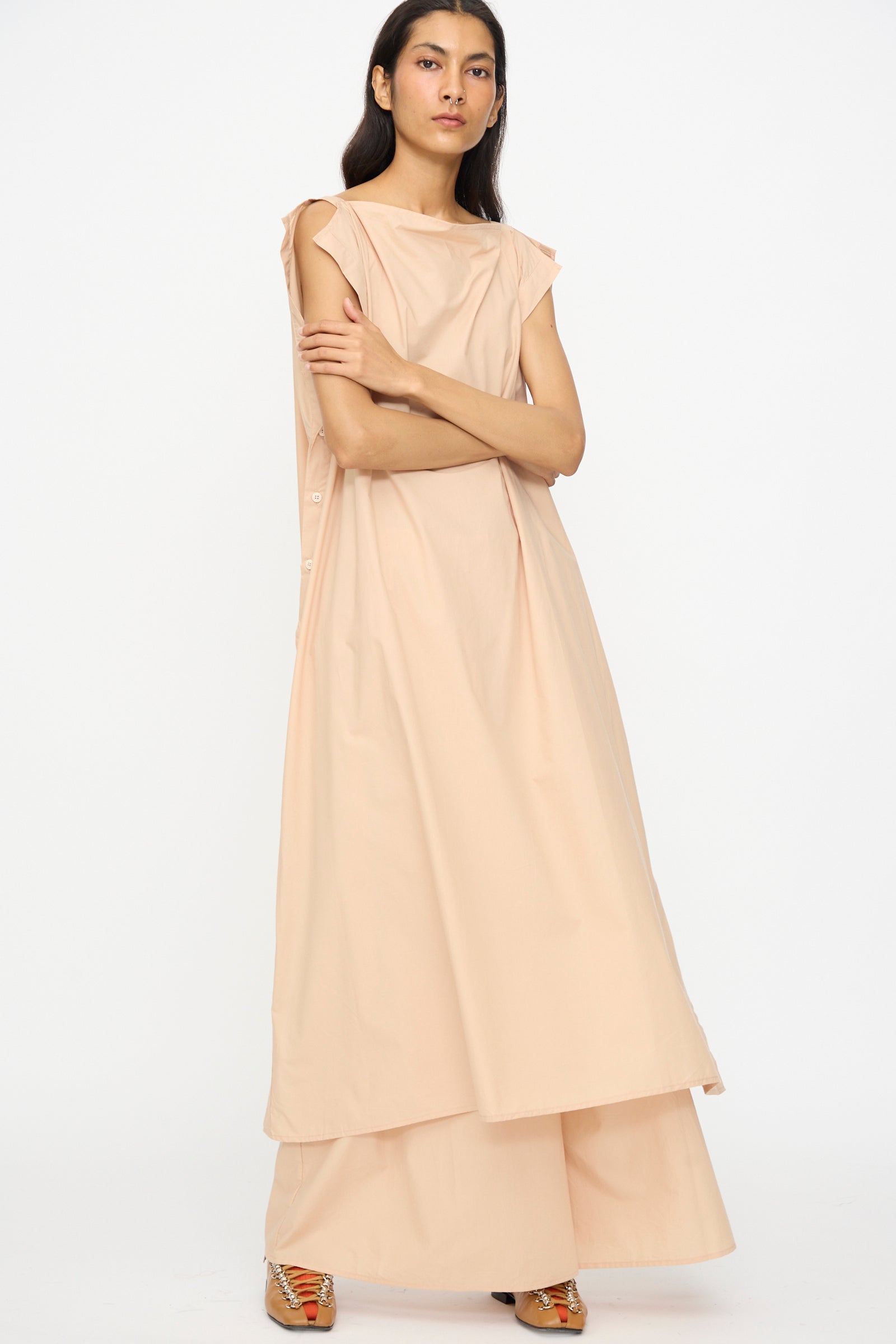 A woman stands with arms crossed, wearing the Baserange Part Dress in Andradita Sand—an oversized fit, sleeveless midi dress—paired with patterned shoes against a plain white background.