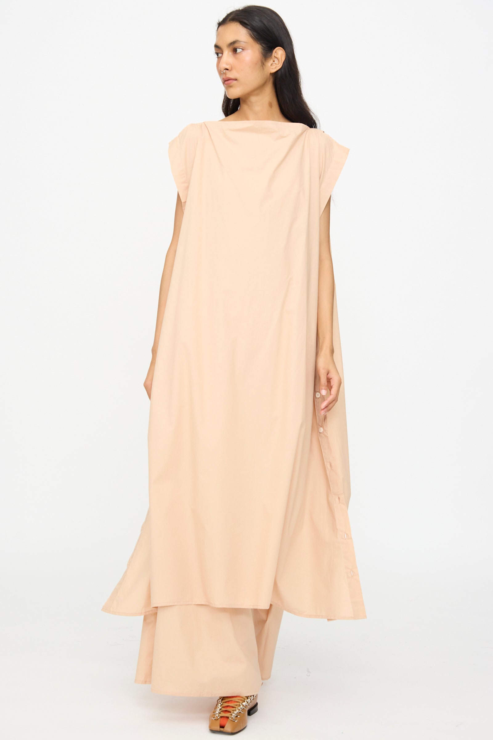 A person wears the Baserange Part Dress in Andradita Sand—an oversized sleeveless midi dress with side buttons—paired with beige wide-leg pants, standing against a plain white background.