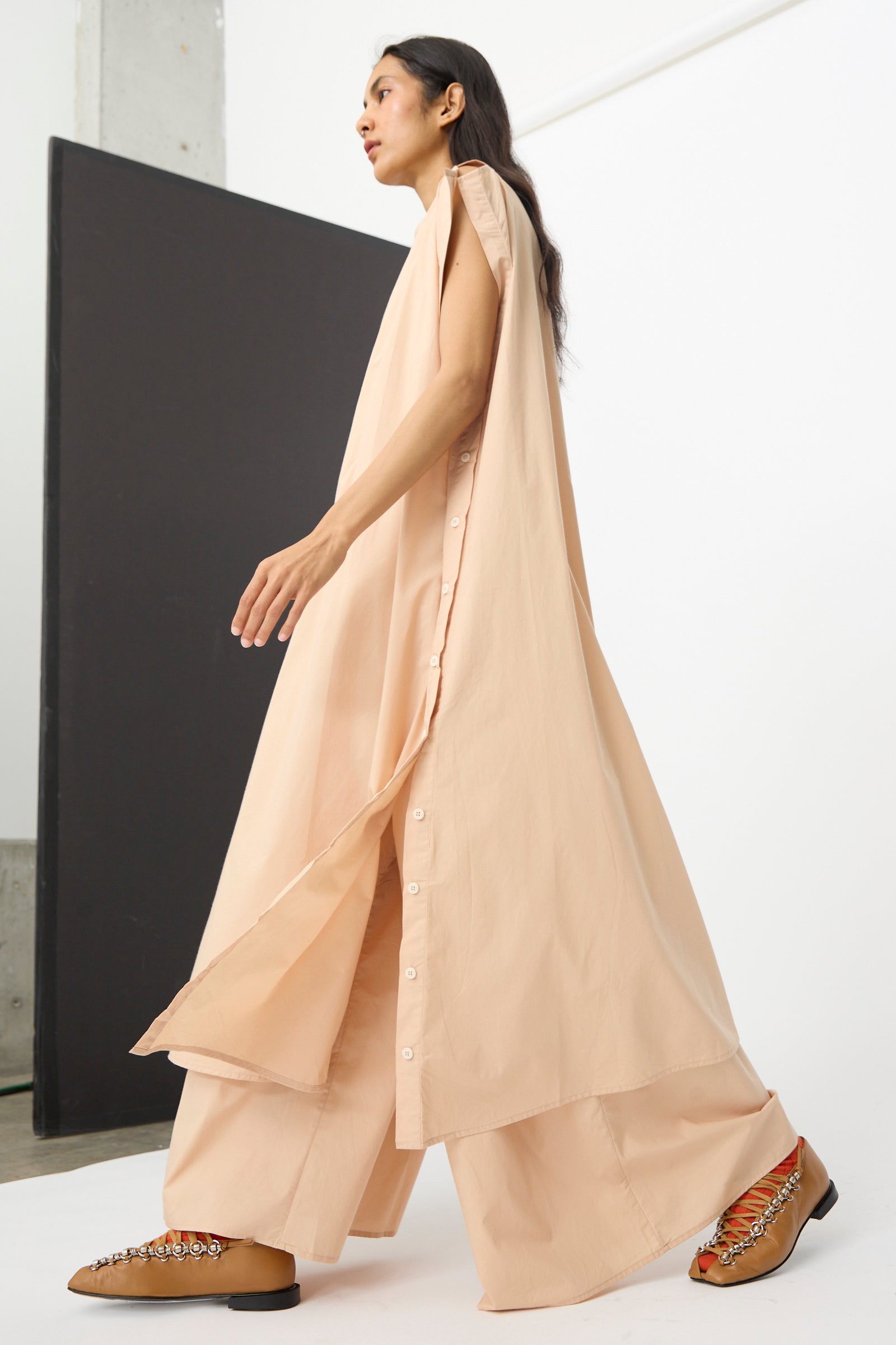 A person wearing the Baserange Part Dress in Andradita Sand styled over wide-legged pants and brown detailed shoes walks indoors.