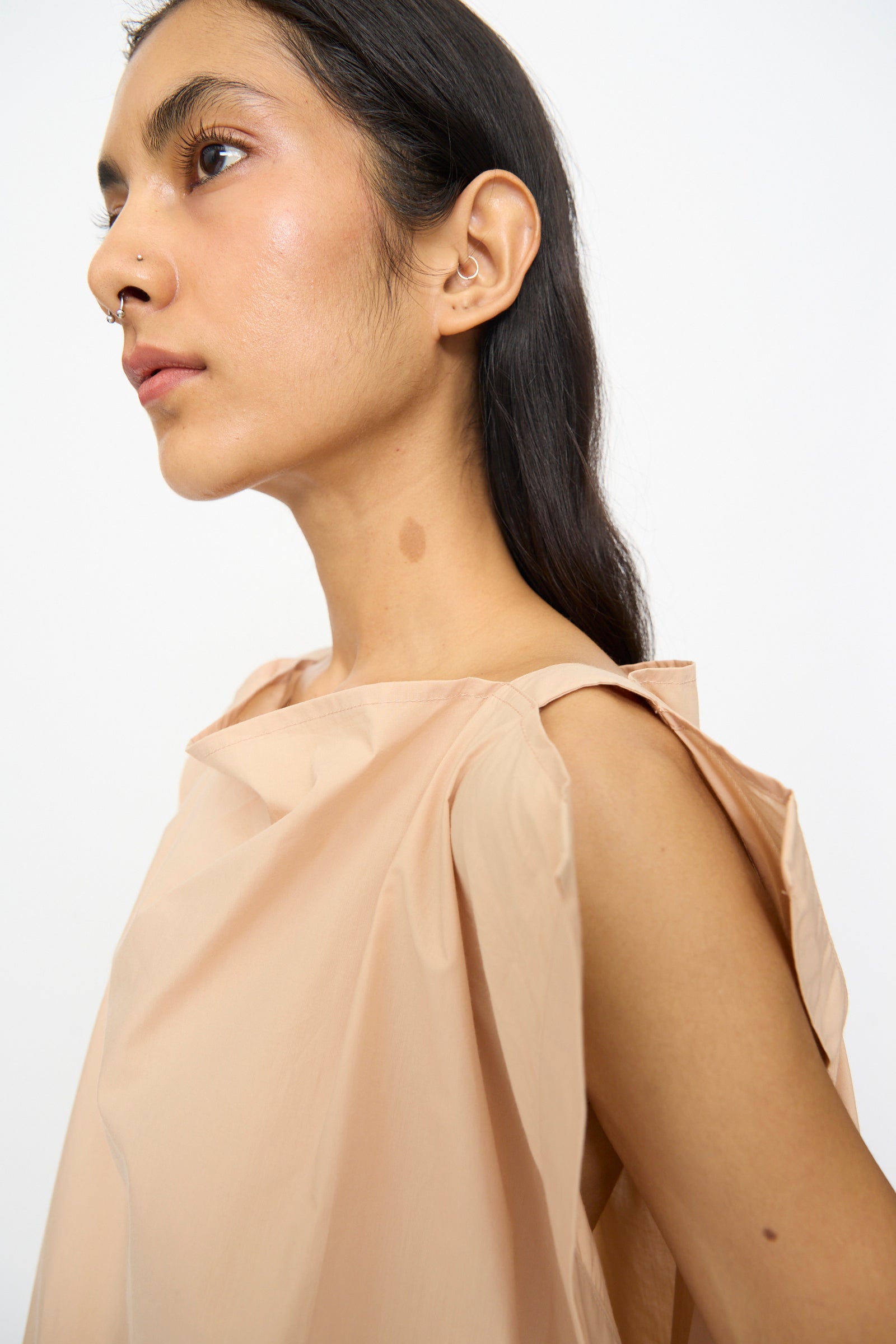 A person with long dark hair and a nose piercing is shown in profile against a plain light background, wearing the Baserange Part Dress in Andradita Sand.