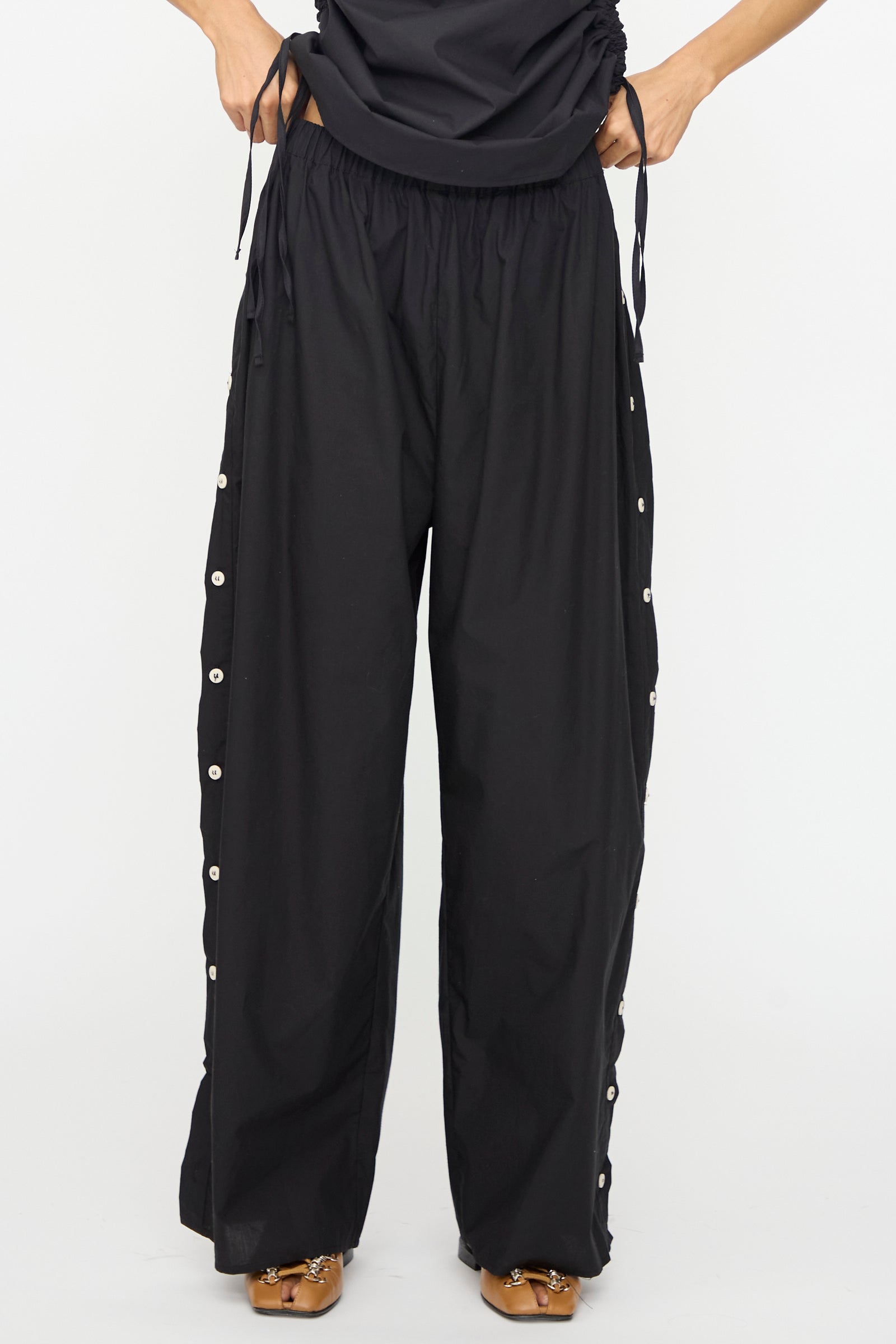 A person wearing Baserange Part Pants in Black with snap buttons down the sides, a black top, and brown sandals stands against a plain white background.