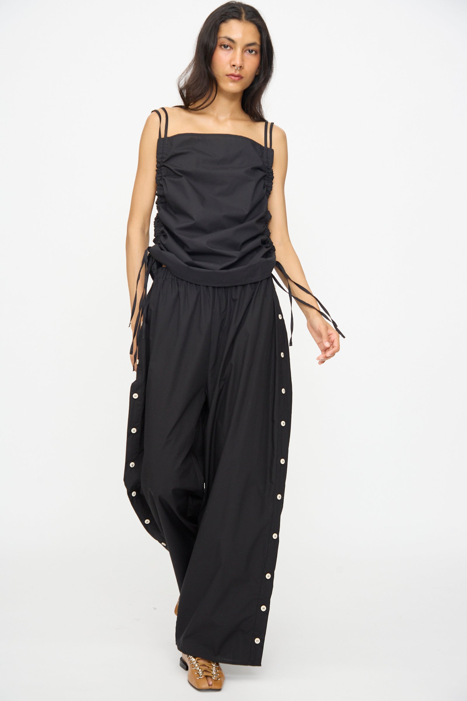 A woman walks forward on a white background wearing Baserange's Part Pants in Black—wide-leg cotton poplin pants with side buttons and an elastic waist—and a black sleeveless ruched top.