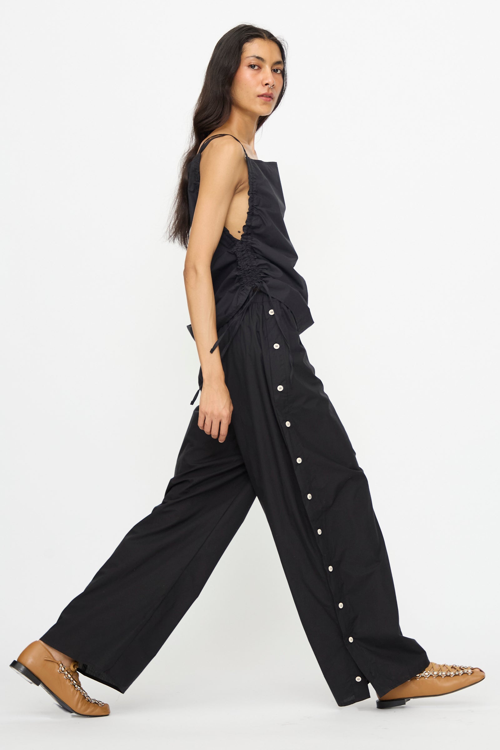 A person walks in profile against a white background, wearing Baserange’s Part Pants in Black—a cotton poplin wide-leg silhouette with side buttons and elastic waist—styled with a sleeveless black top and tan shoes.