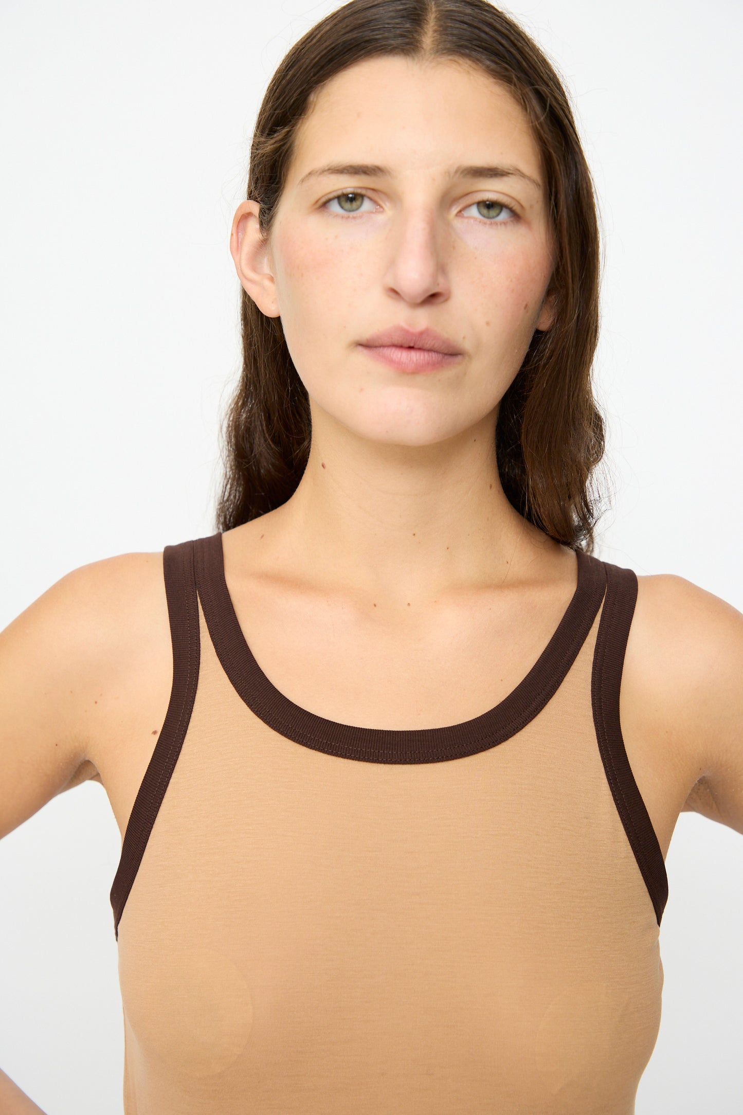 A person with long brown hair wears the Baserange Racer Tank in Aural Tactile, a slim fit tank top with dark brown trim made in Portugal, standing against a plain white background.