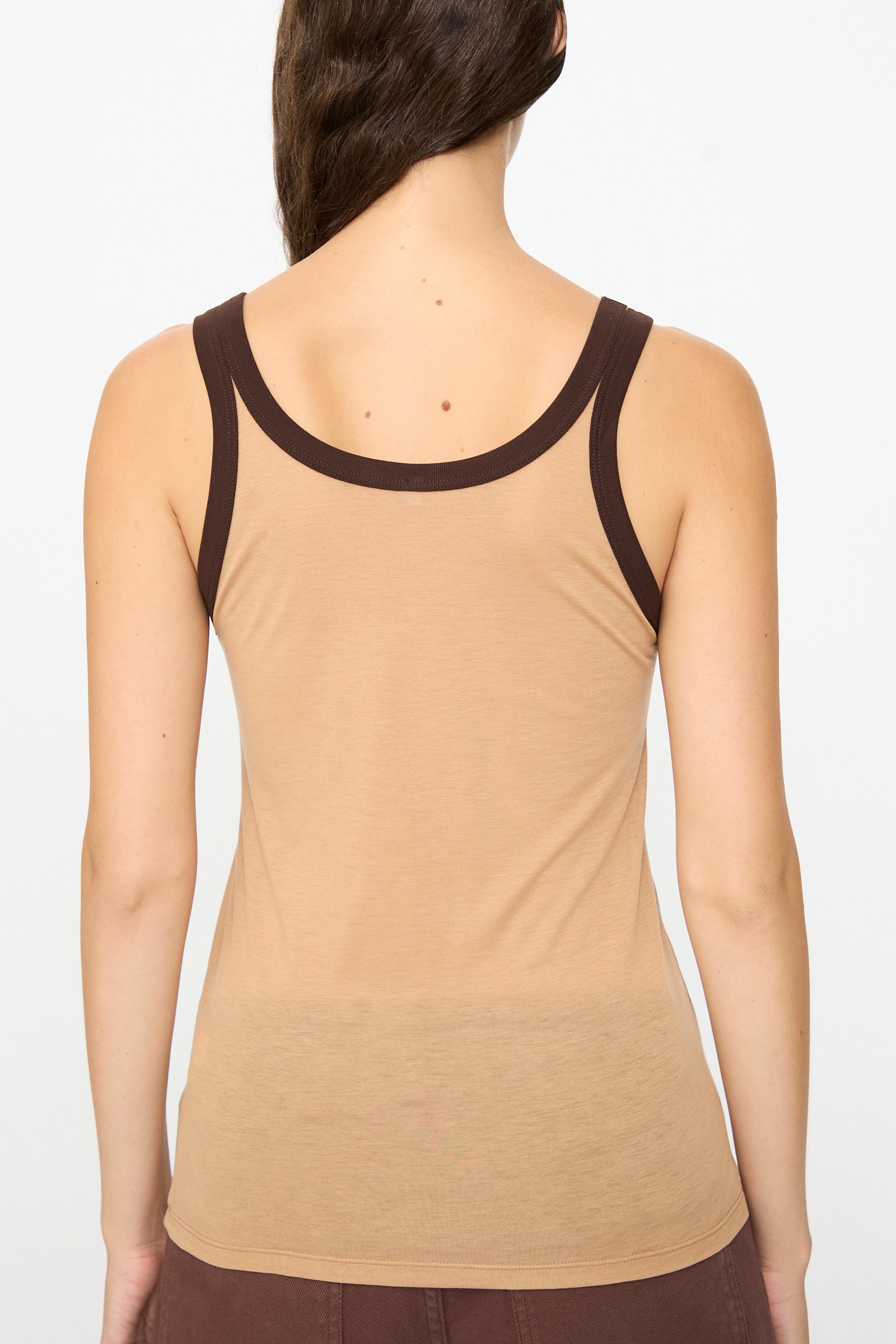 A person with long brown hair facing away, wearing the Baserange Racer Tank in Aural Tactile—a beige bamboo lyocell tank top with dark brown trim—paired with brown pants, stands against a plain white background.
