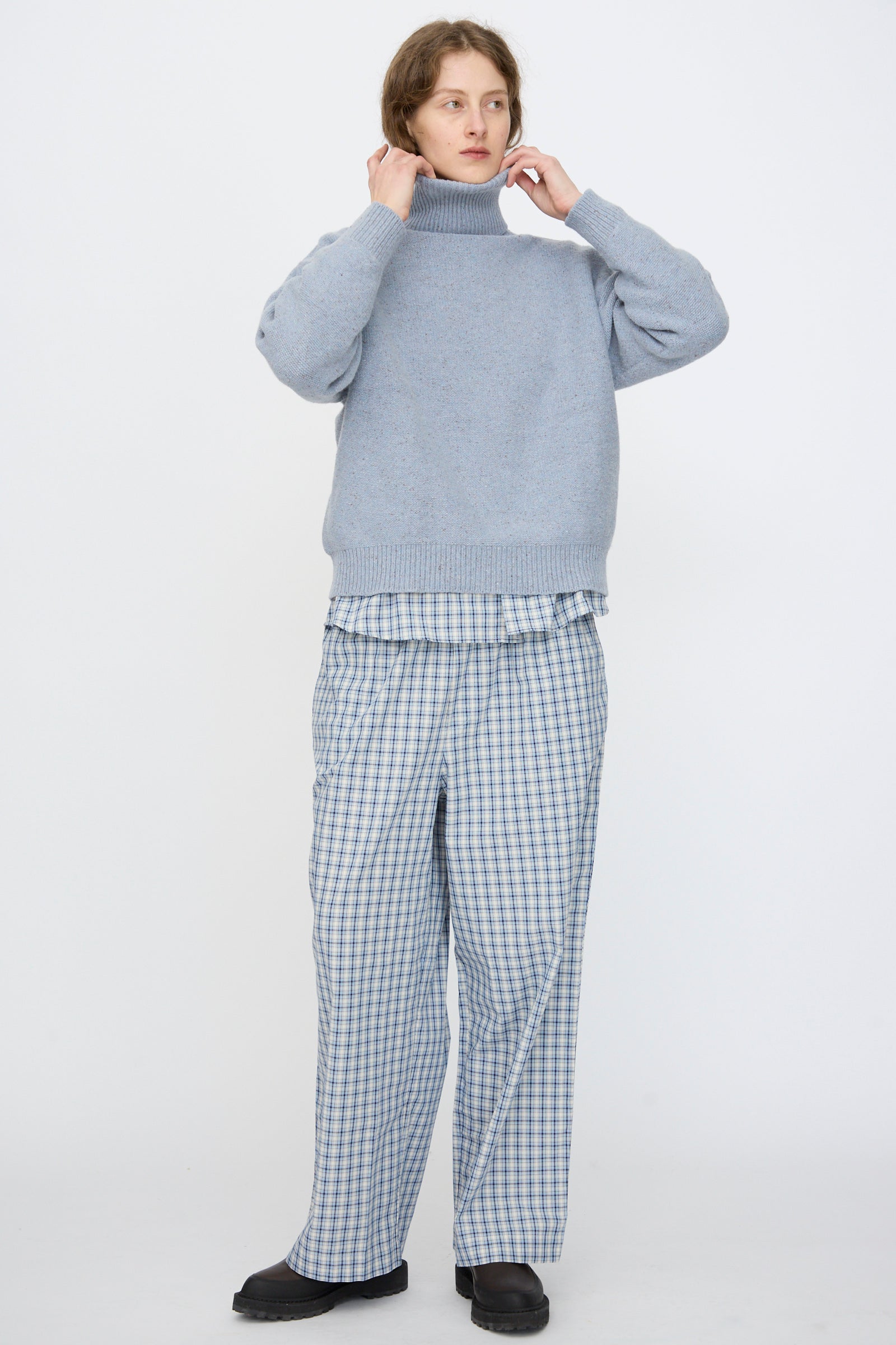 A person stands against a plain background wearing the Baserange Recycled Wool Mea Turtleneck in Ocean, paired with loose plaid pants and black shoes. 