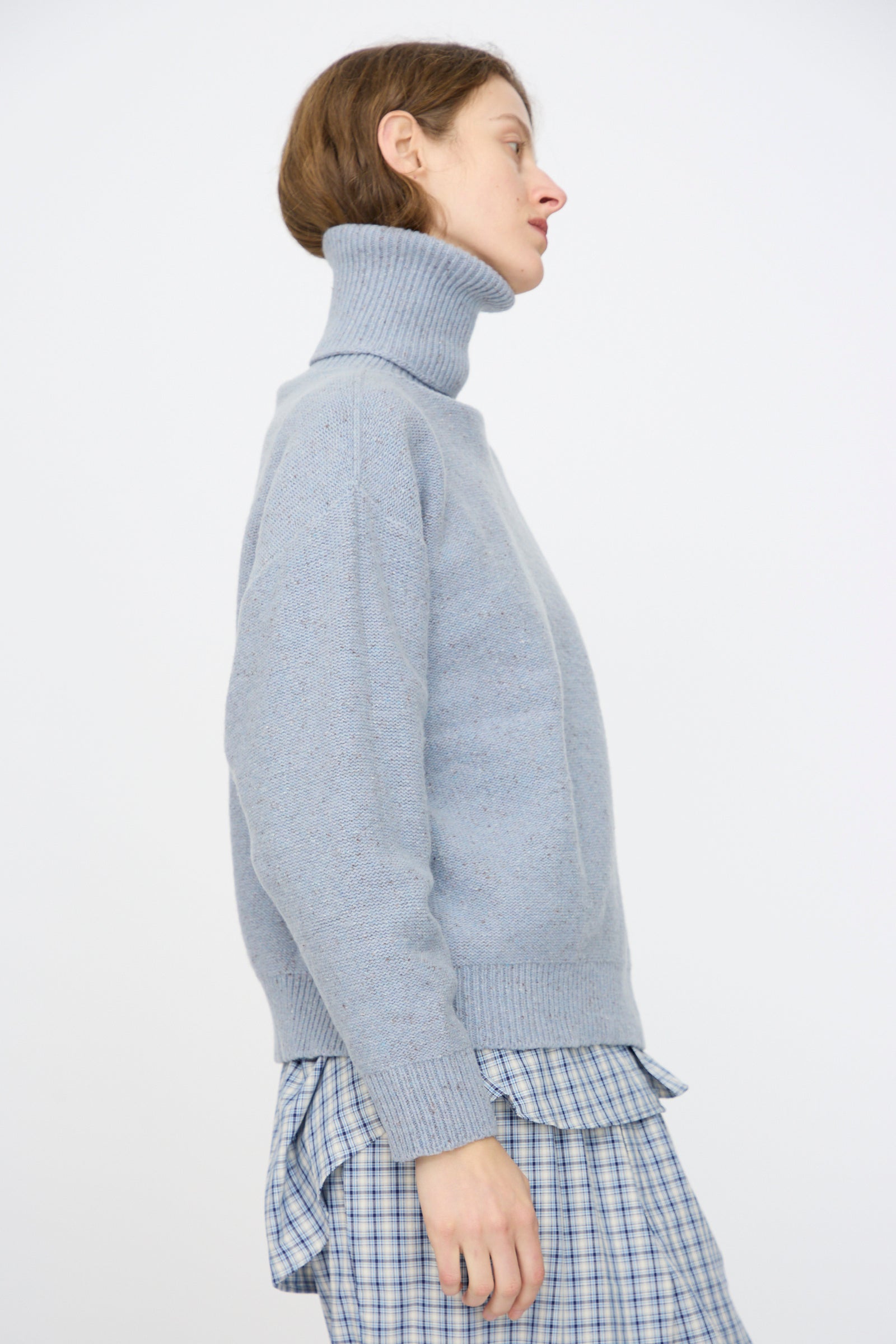 A person in profile wears the Baserange Recycled Wool Mea Turtleneck in Ocean layered over a blue and white plaid dress, standing against a plain white background.