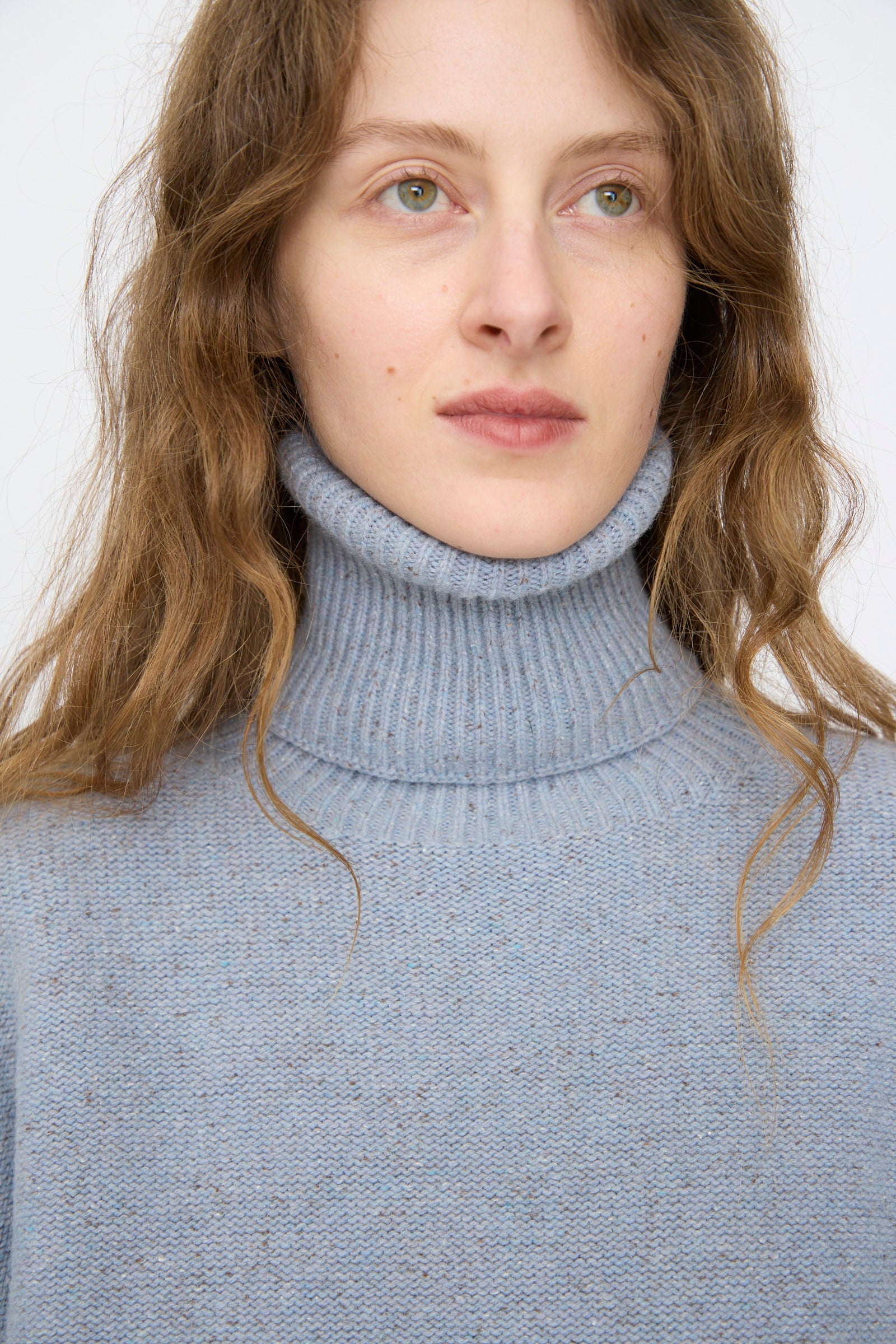 A person with long brown hair wears the Baserange Recycled Wool Mea Turtleneck in Ocean, looking slightly to the side against a plain background.