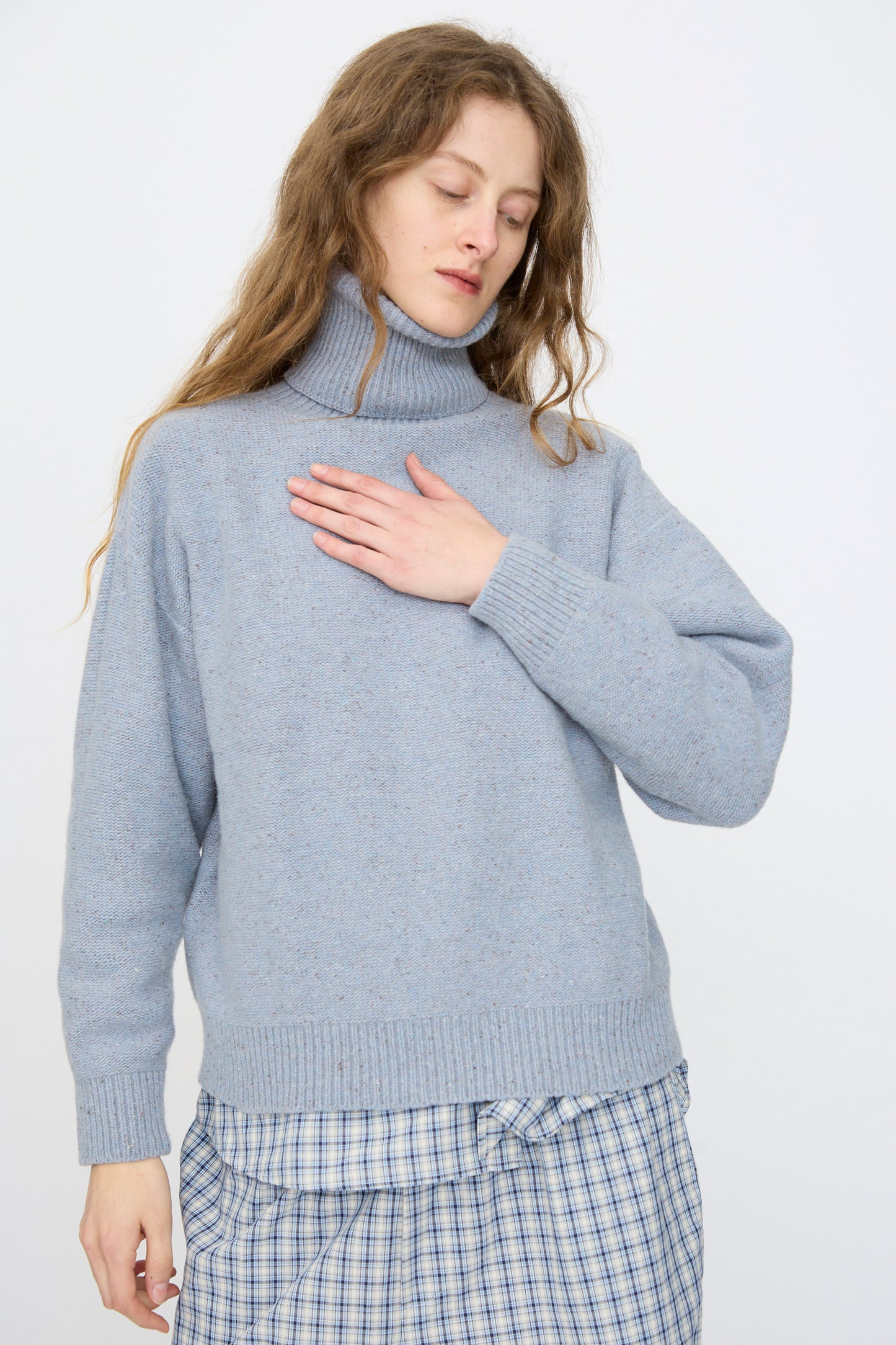 A person with long hair wears the Baserange Recycled Wool Mea Turtleneck in Ocean, paired with a plaid skirt, standing with one hand on their chest and looking down.