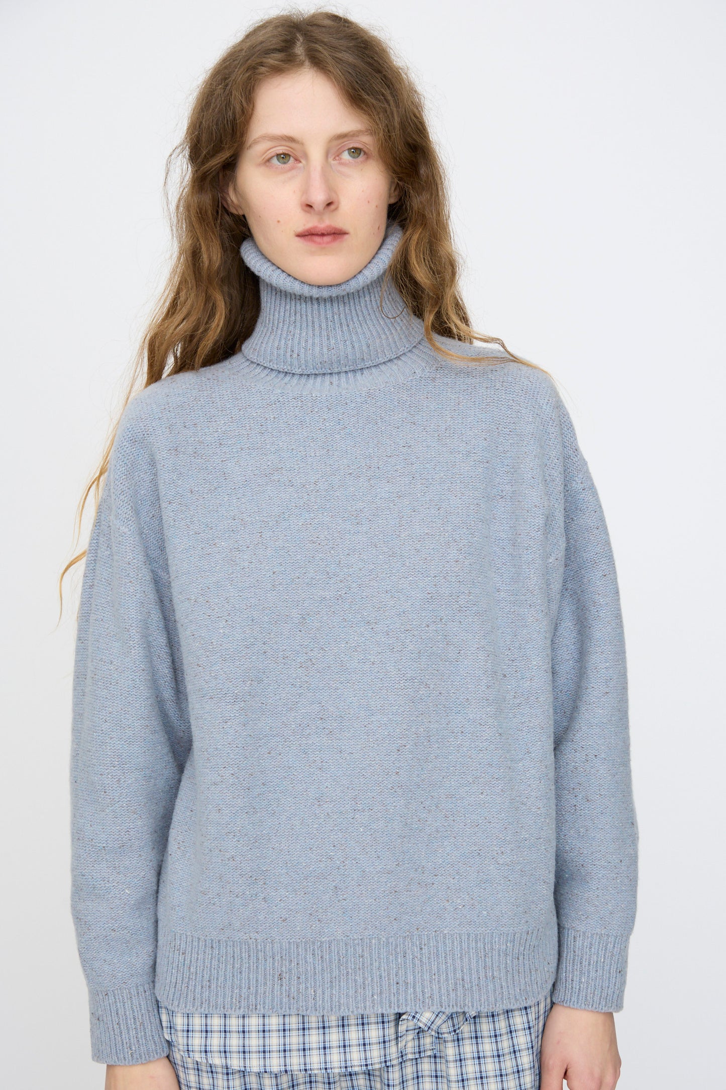A person with long hair wears the Baserange Recycled Wool Mea Turtleneck in Ocean, paired with blue plaid pants, standing against a plain light background.