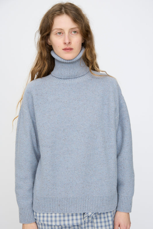 A person with long hair wears the Baserange Recycled Wool Mea Turtleneck in Ocean, paired with blue plaid pants, standing against a plain light background.